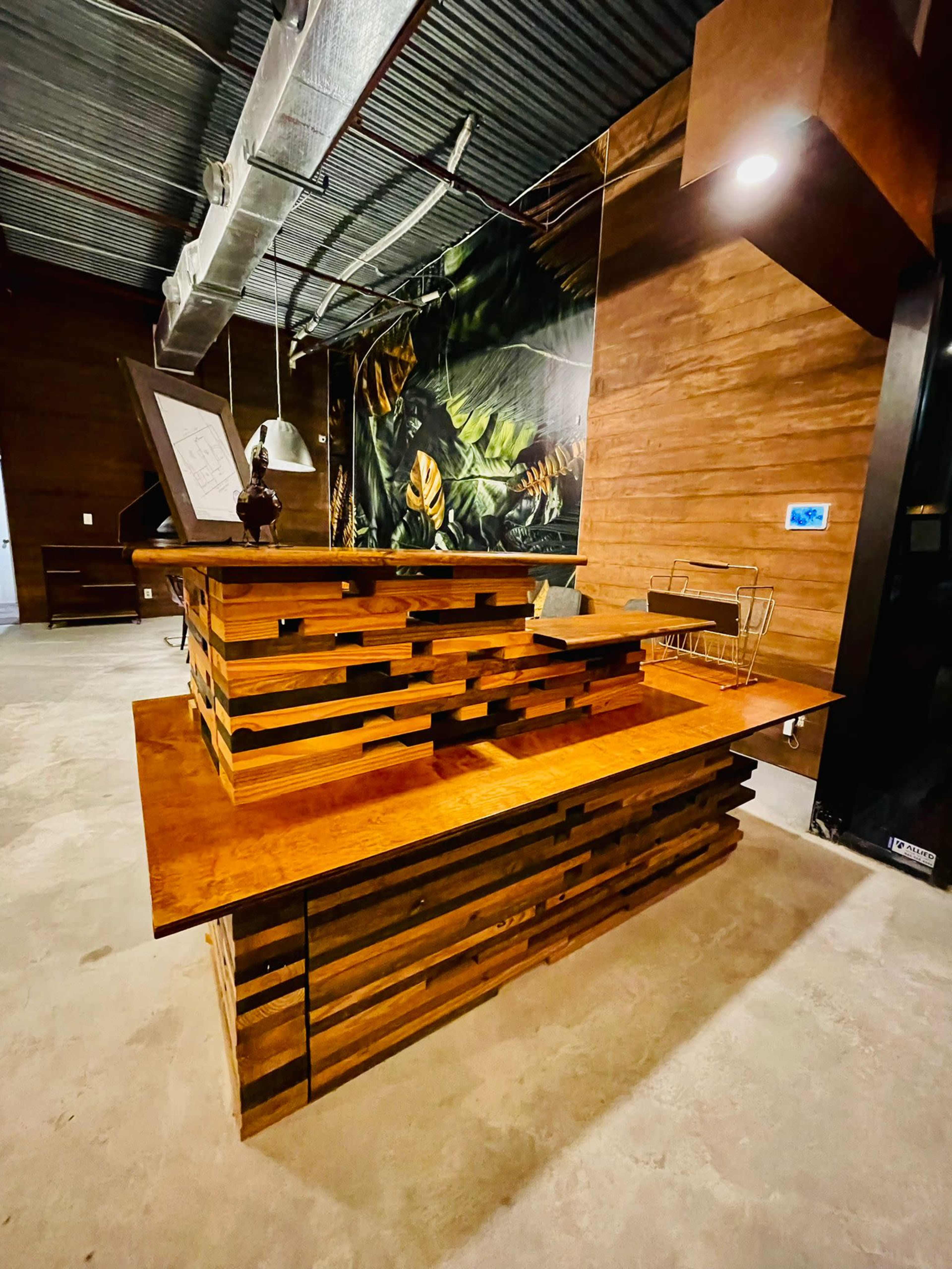 A modern wooden reception desk made of layered planks, situated in an industrial-style interior with a mural of tropical leaves in the background.