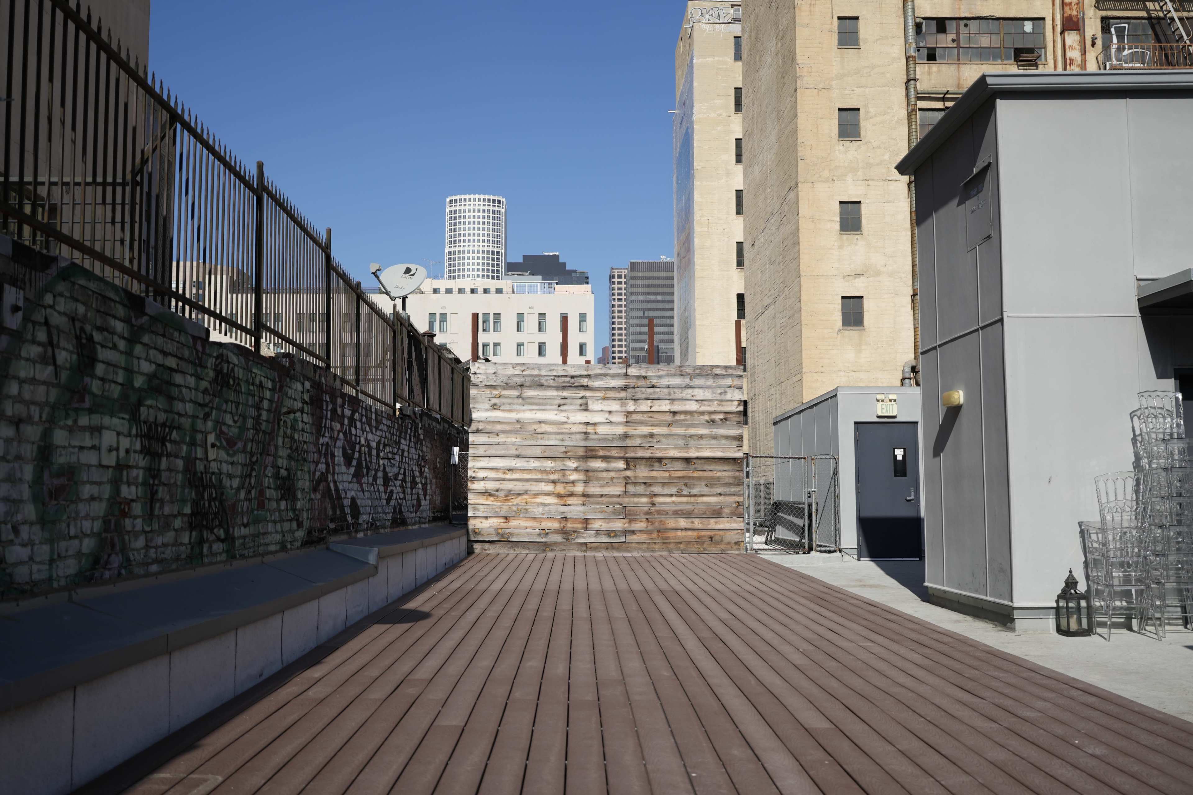 The image shows a rooftop space with wooden decking, a wooden wall in the background, and a city skyline visible beyond a fence.