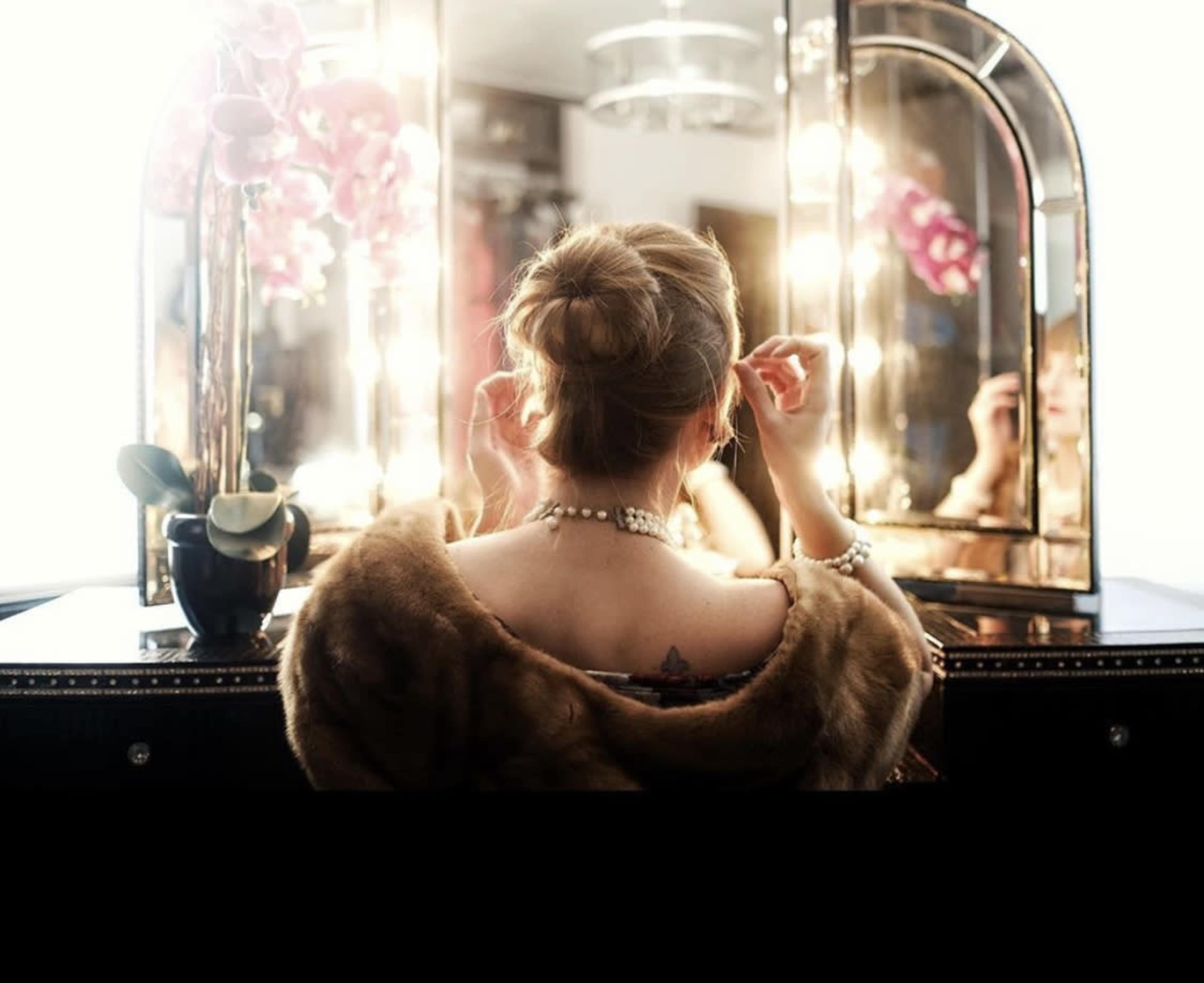 A woman in a fur wrap is seated in front of a mirrored vanity, adjusting her hair under soft, glowing lights.