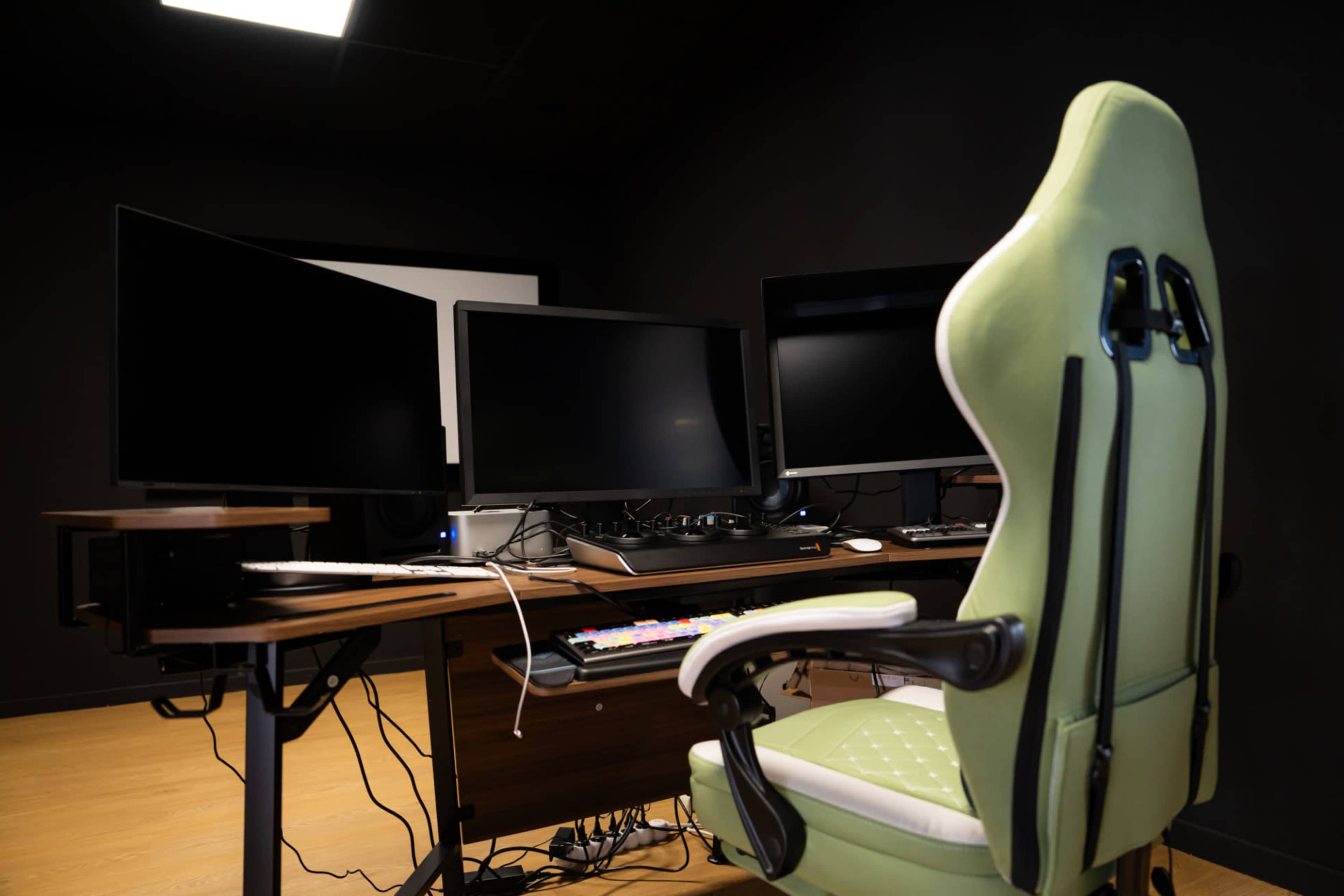 A green gaming chair is positioned in front of a wooden desk with multiple monitors and a keyboard, in a dark room.