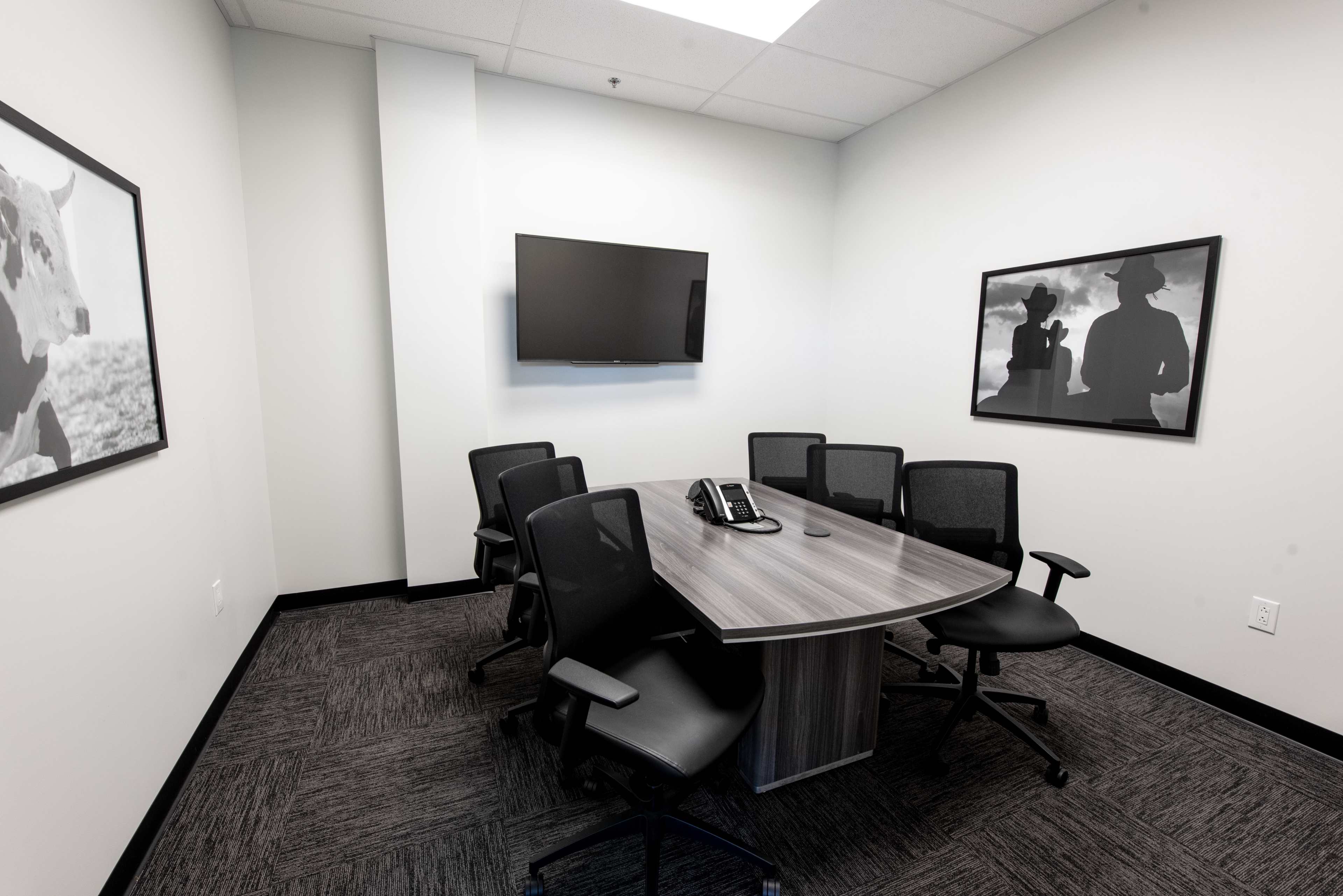 A modern conference room with a round table, six chairs, a television mounted on the wall, and black-and-white artwork framing the space.