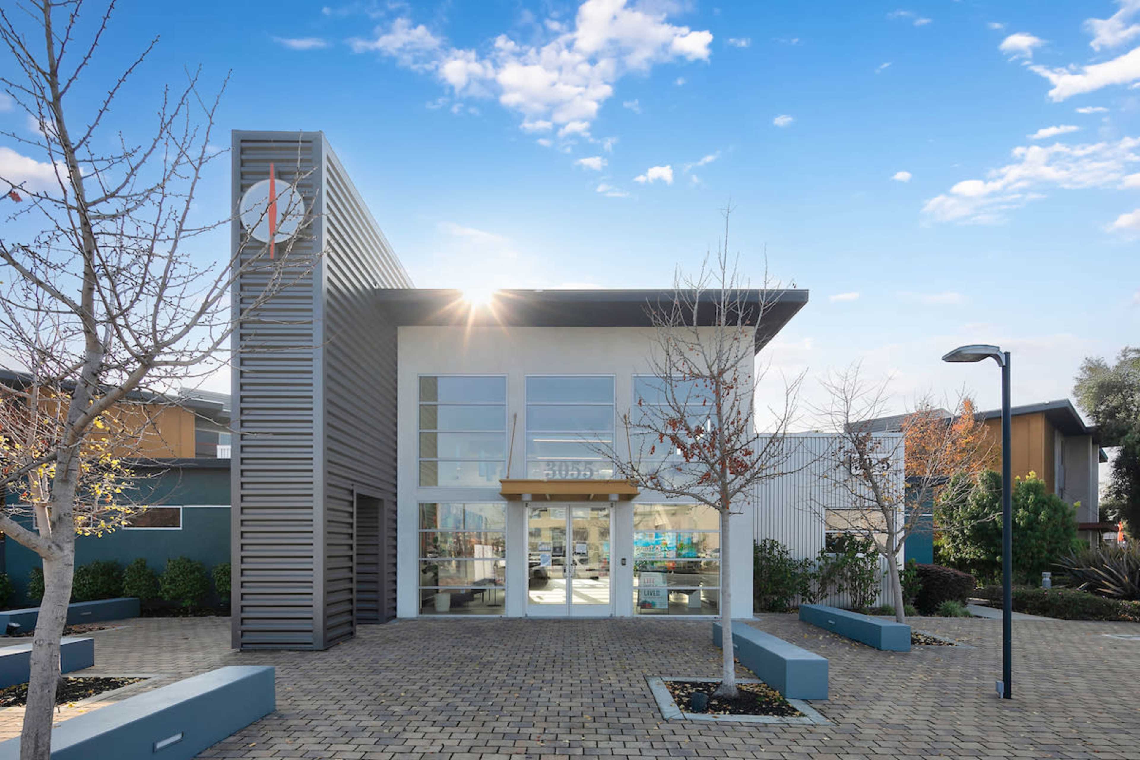 A modern building with a grid-paved walkway, large glass windows, and a prominent sign featuring a logo, surrounded by leafless trees and landscaped greenery.