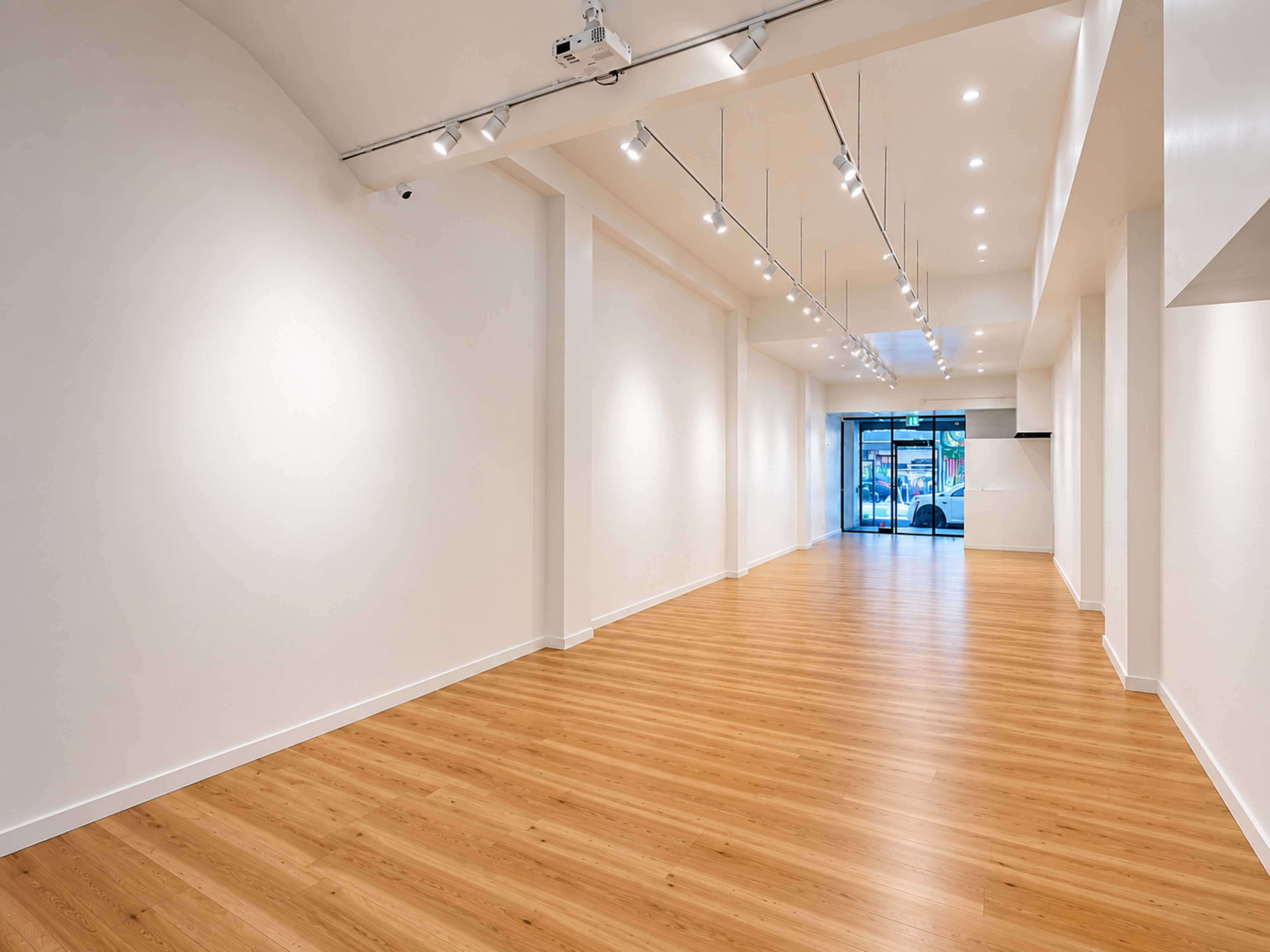 The image shows a spacious, modern interior with wooden flooring and white walls, illuminated by track lighting and large windows at the entrance.