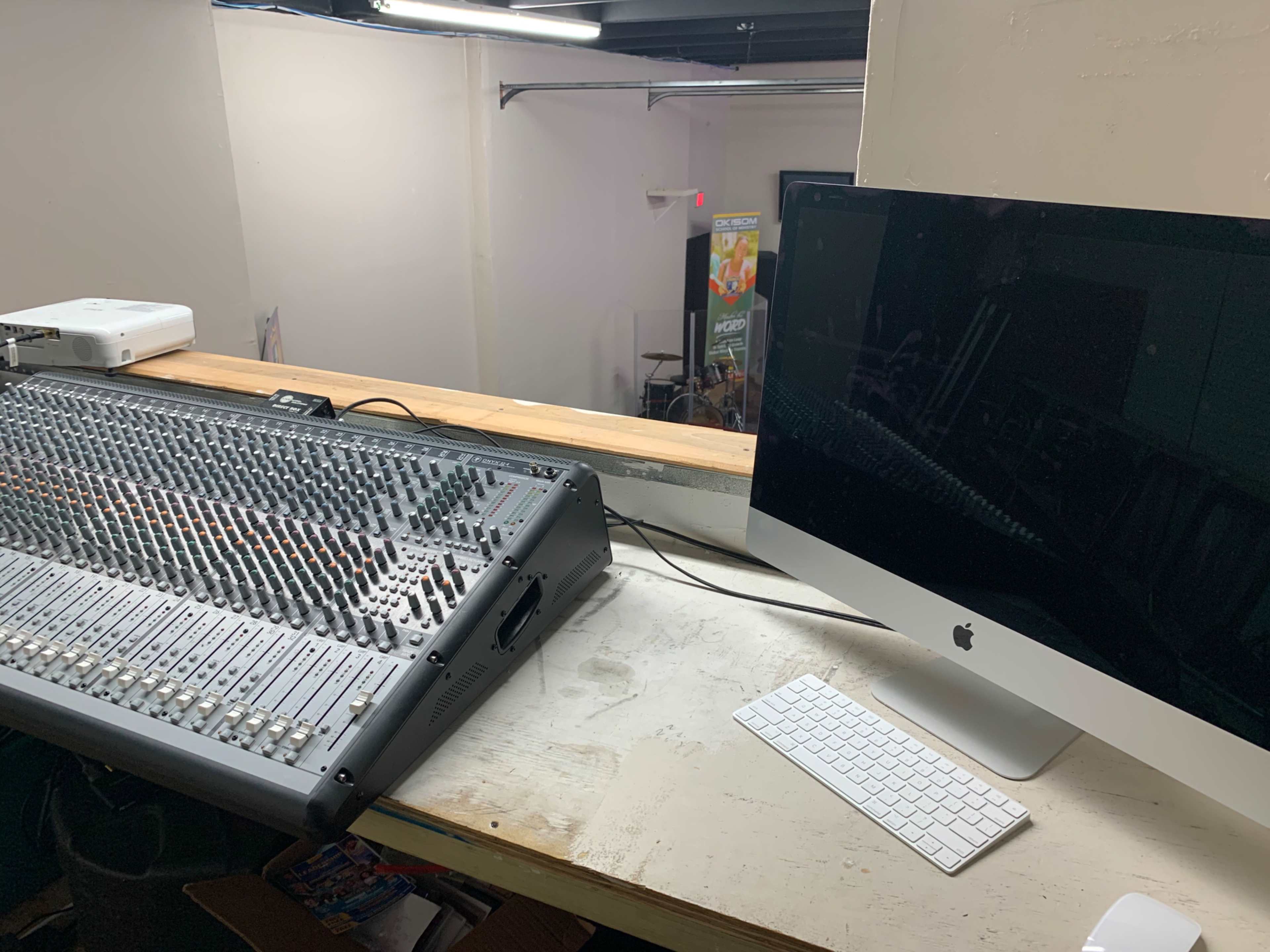 A sound mixing console is positioned next to an iMac computer on a wooden table in a studio space.