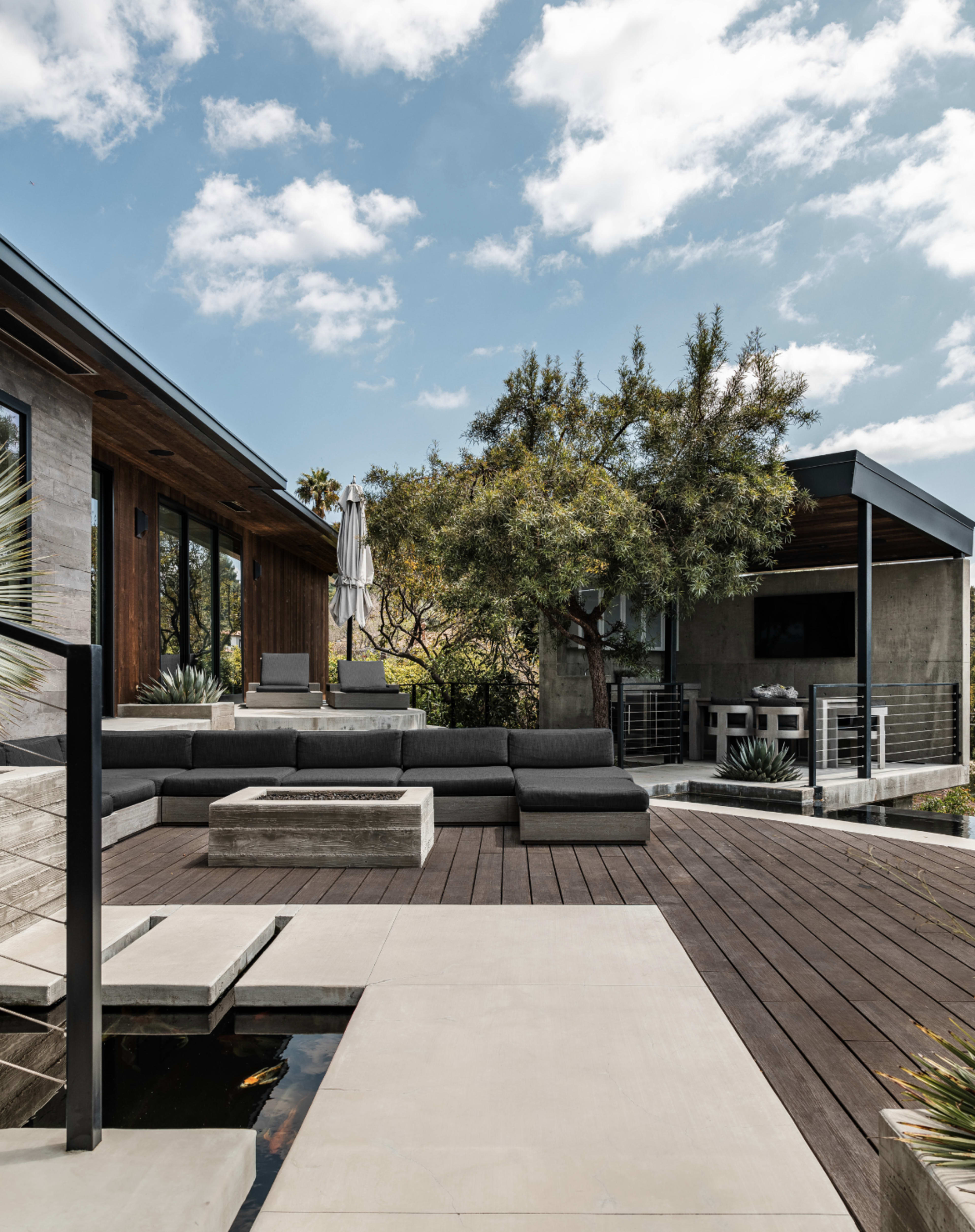 A modern outdoor living space features a large sectional sofa, a fire pit, and a shaded area with a table, surrounded by greenery and blue skies.