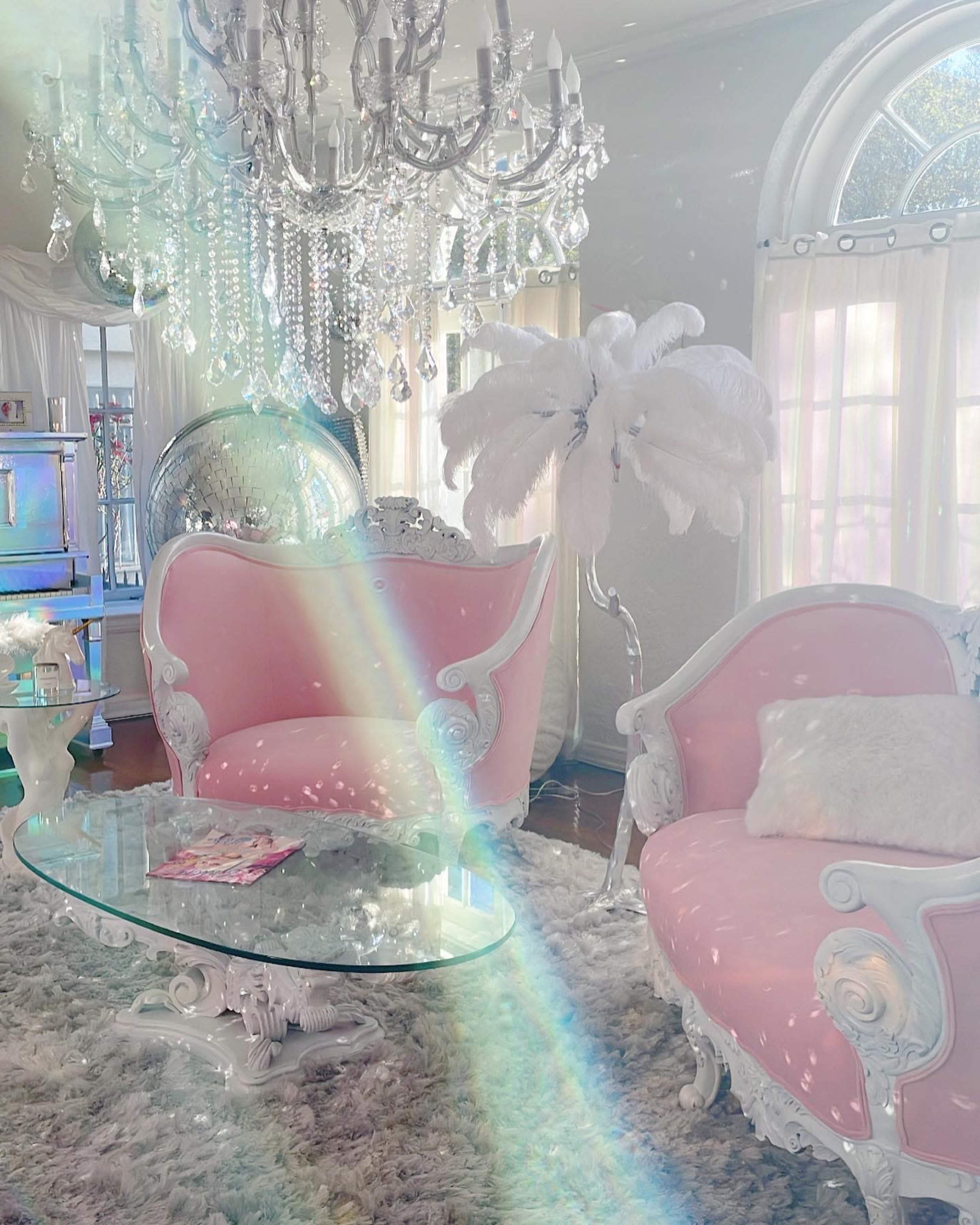 A bright sitting area with pink upholstered chairs, a glass coffee table, a chandelier, and a decorative feathered floor lamp.
