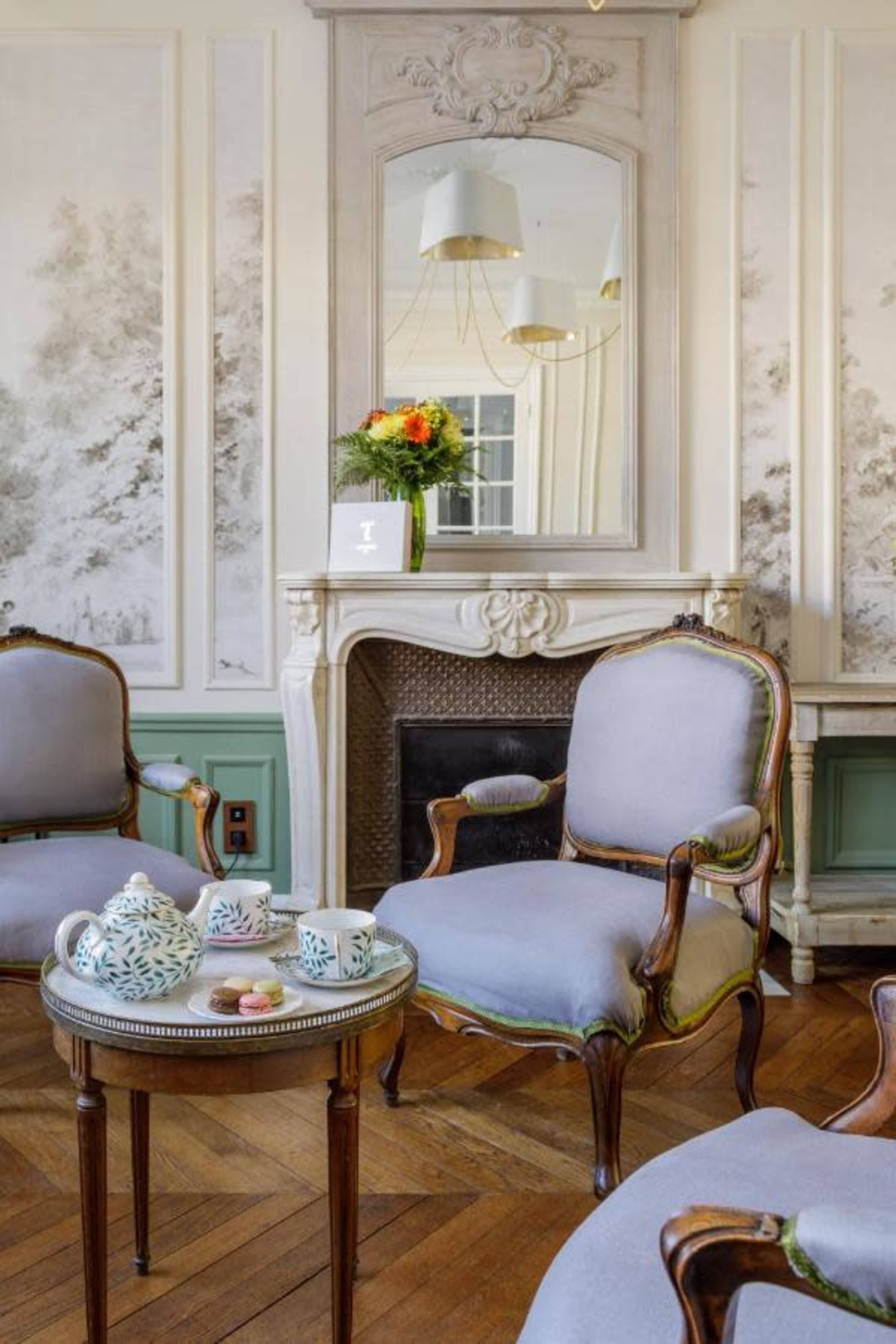 A stylish sitting area featuring gray upholstered chairs, a round wooden table with decorative teacups, and a fireplace adorned with a floral arrangement and a large mirror.
