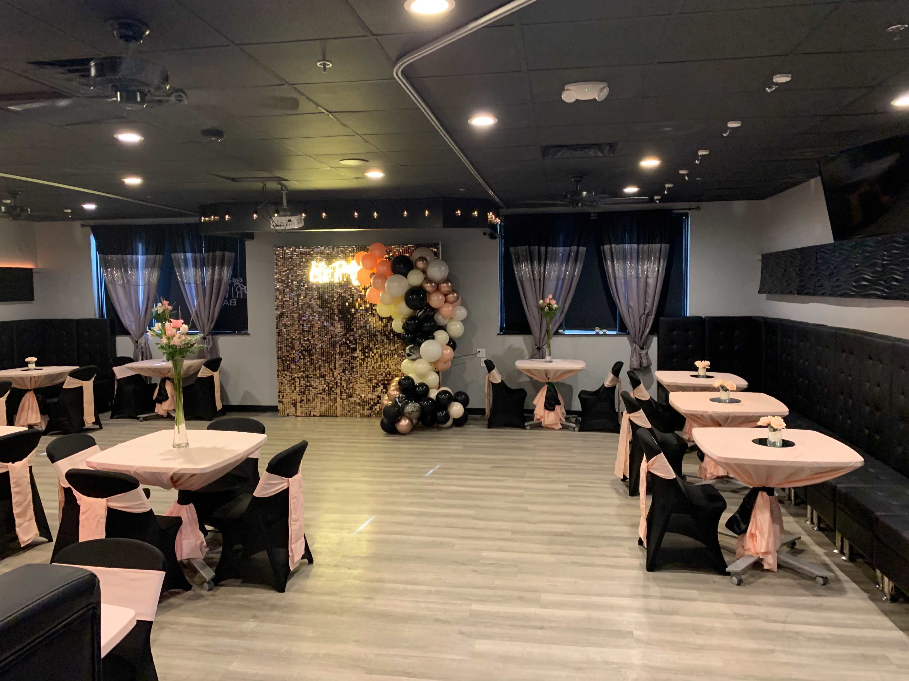 A decorated event space with tables covered in black and pink cloths, surrounded by black chairs, and features a balloon backdrop.