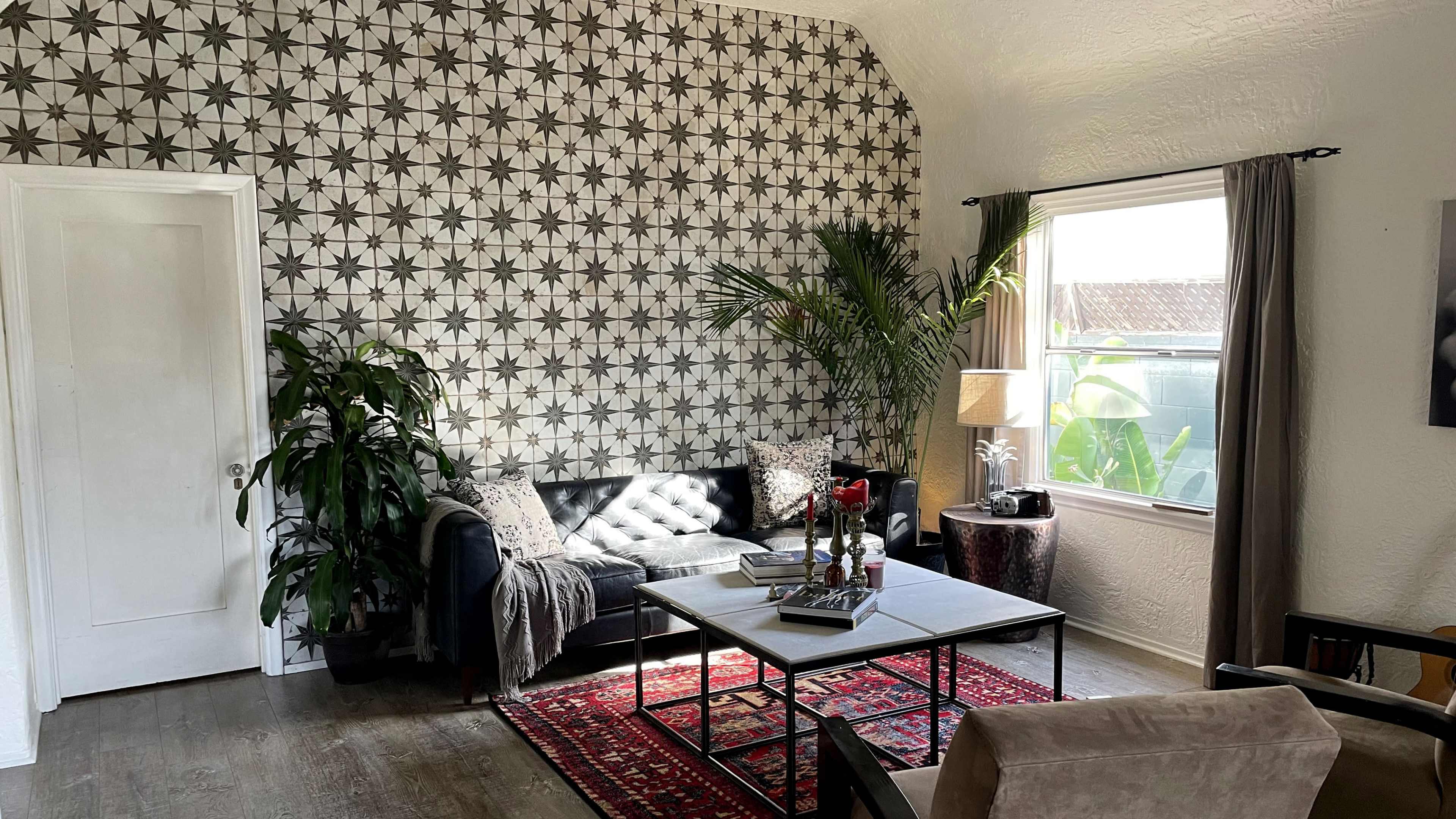 The image shows a living room with a patterned wall, a black sofa adorned with a blanket, a coffee table with decor, and a potted plant beside a window.