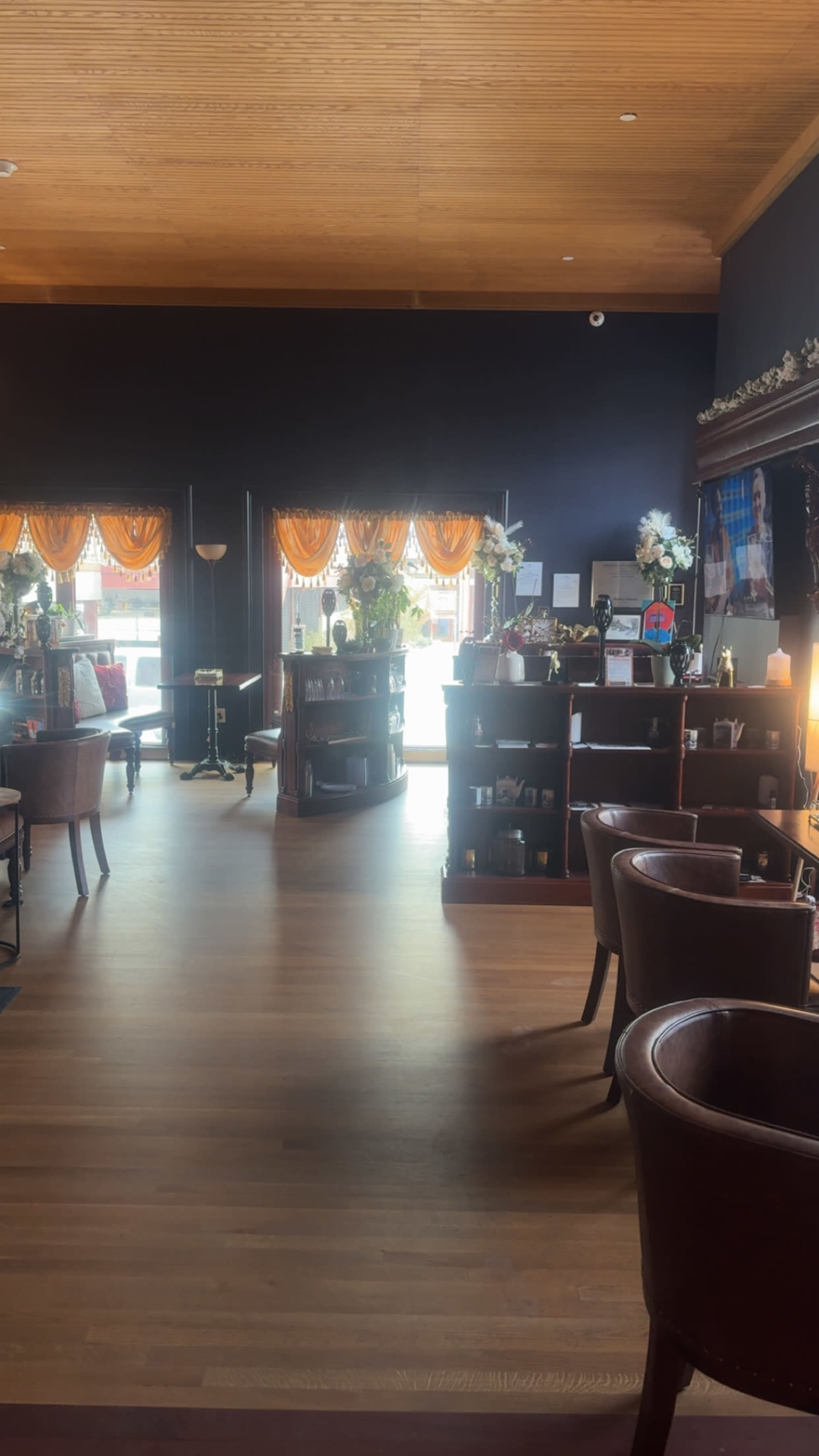 The image shows the interior of a café with wooden flooring, shelves filled with items, and decorative curtains framing large windows.