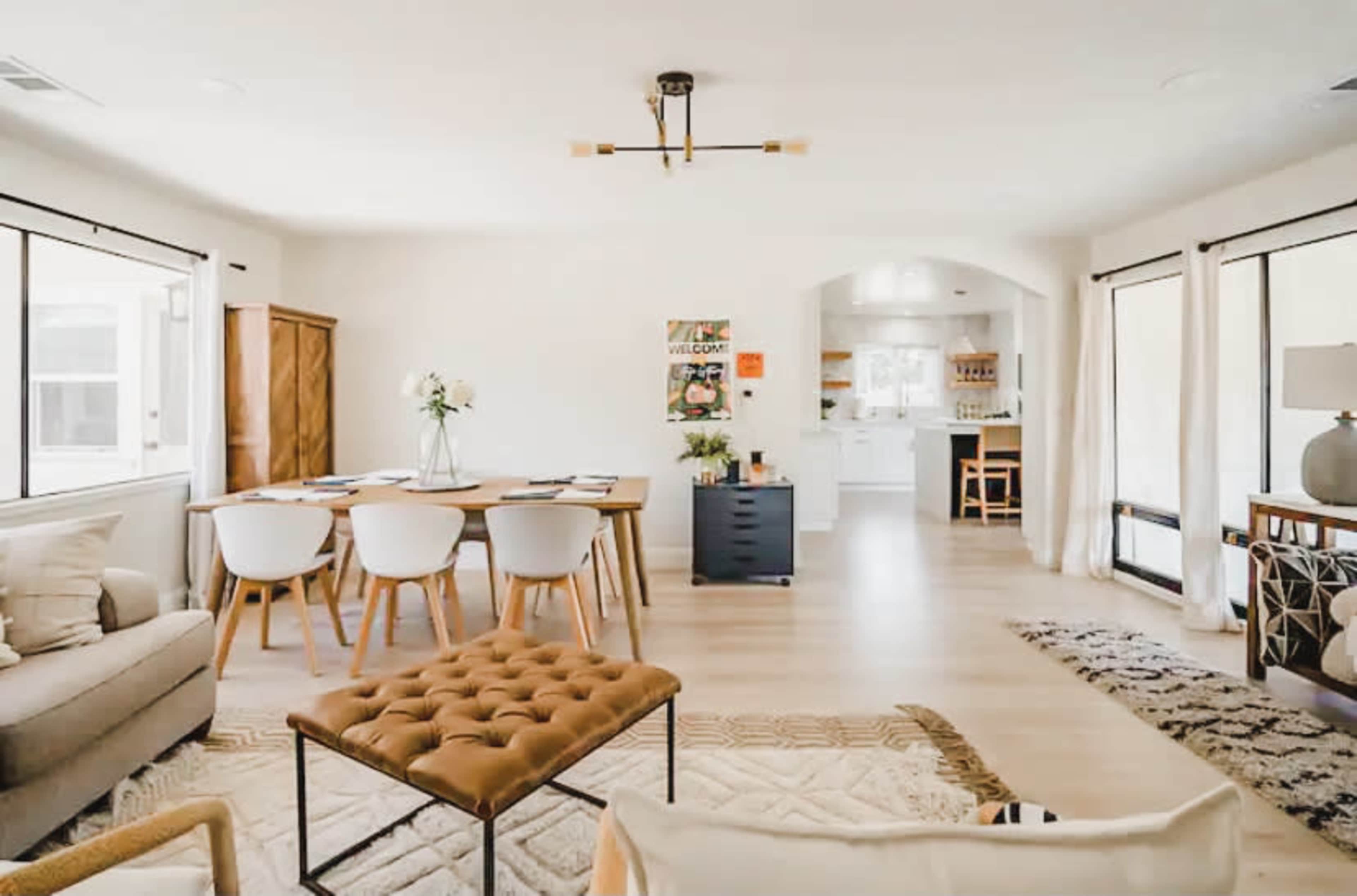 A bright, open living space with a dining area featuring a wooden table and white chairs, leading to a kitchen in the background.