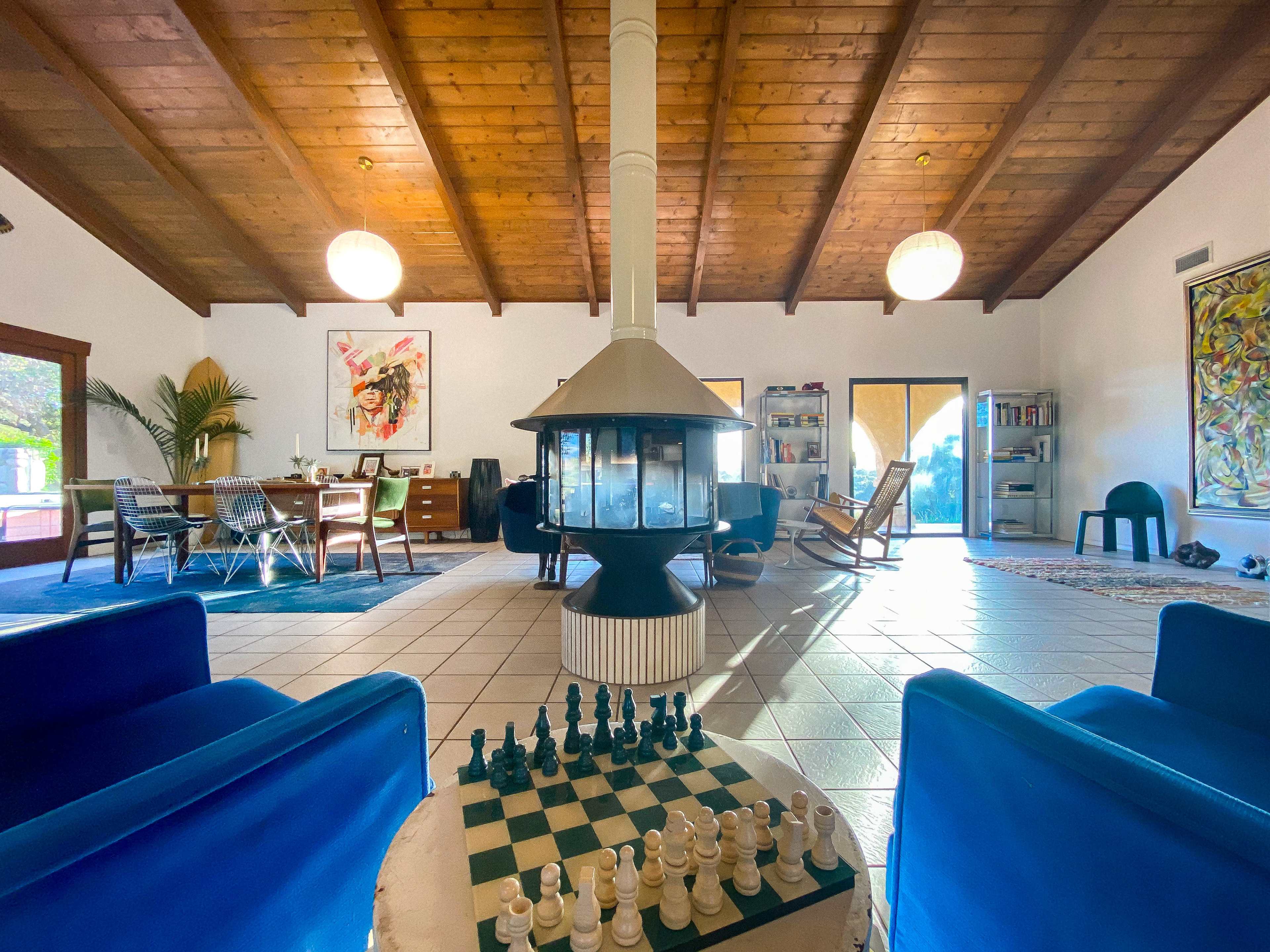 The image shows a spacious living area with a chess set, a central fireplace, wooden beams in the ceiling, and a mix of modern and traditional furniture.