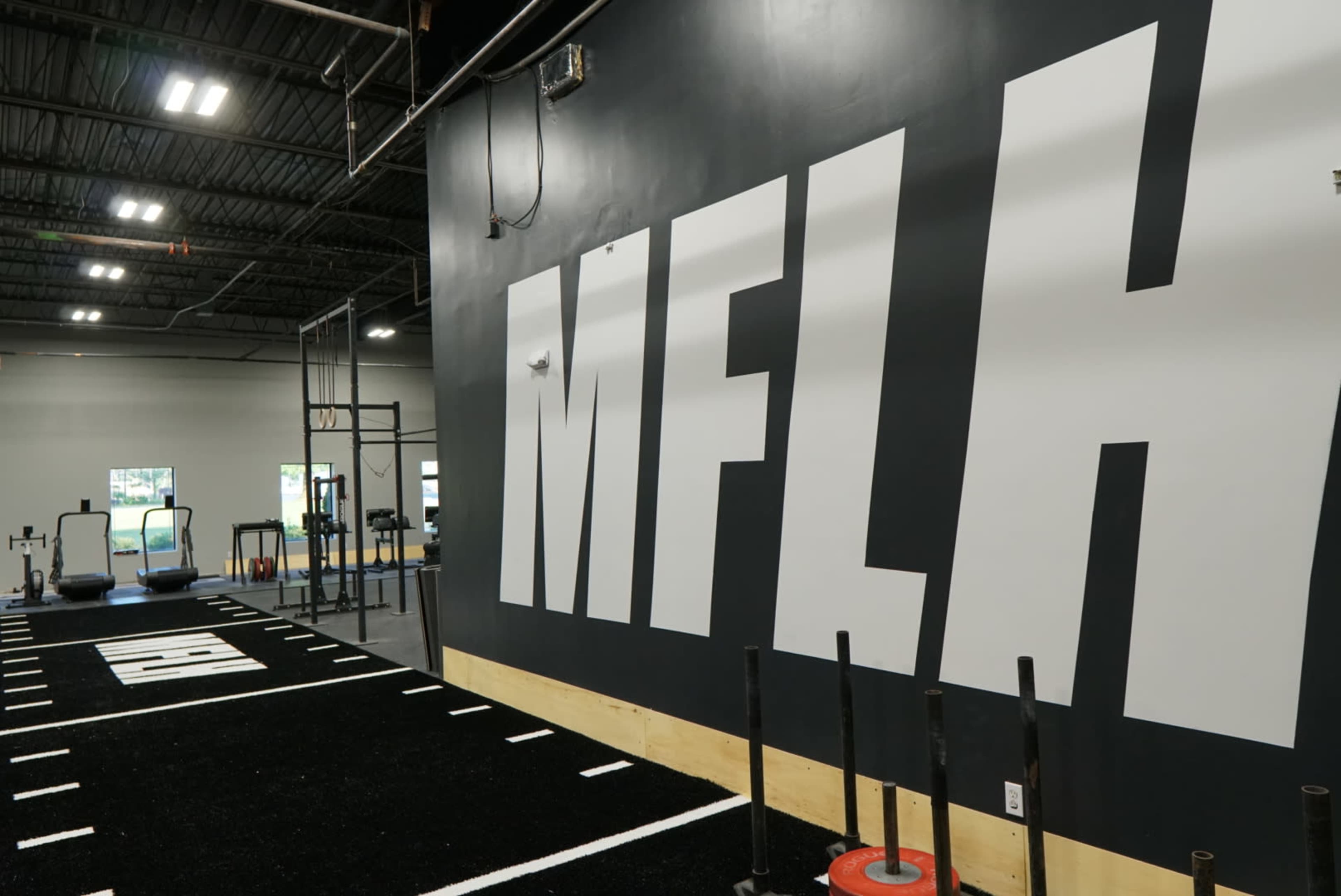 The image shows a fitness training facility with a large "MFLH" logo on the wall and workout equipment arranged along a turf-lined area.