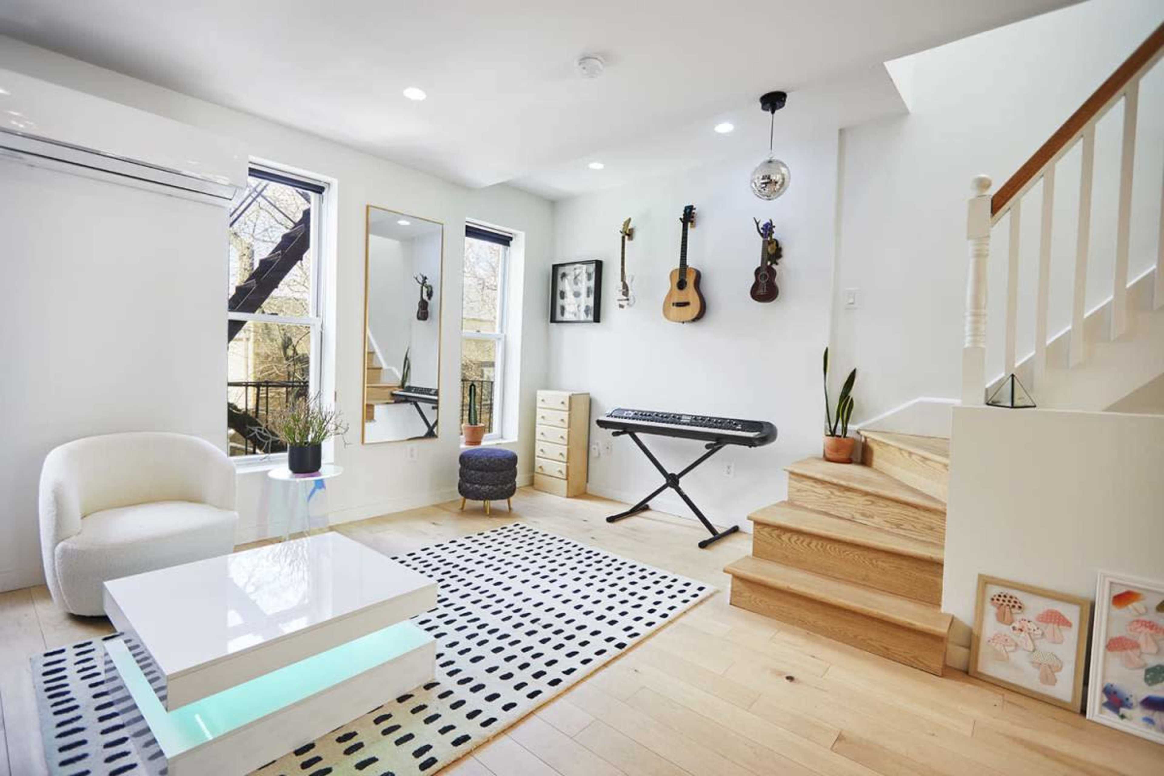 The image shows a modern living room with light wood flooring, a white coffee table, a keyboard, and a staircase leading to an upper level.