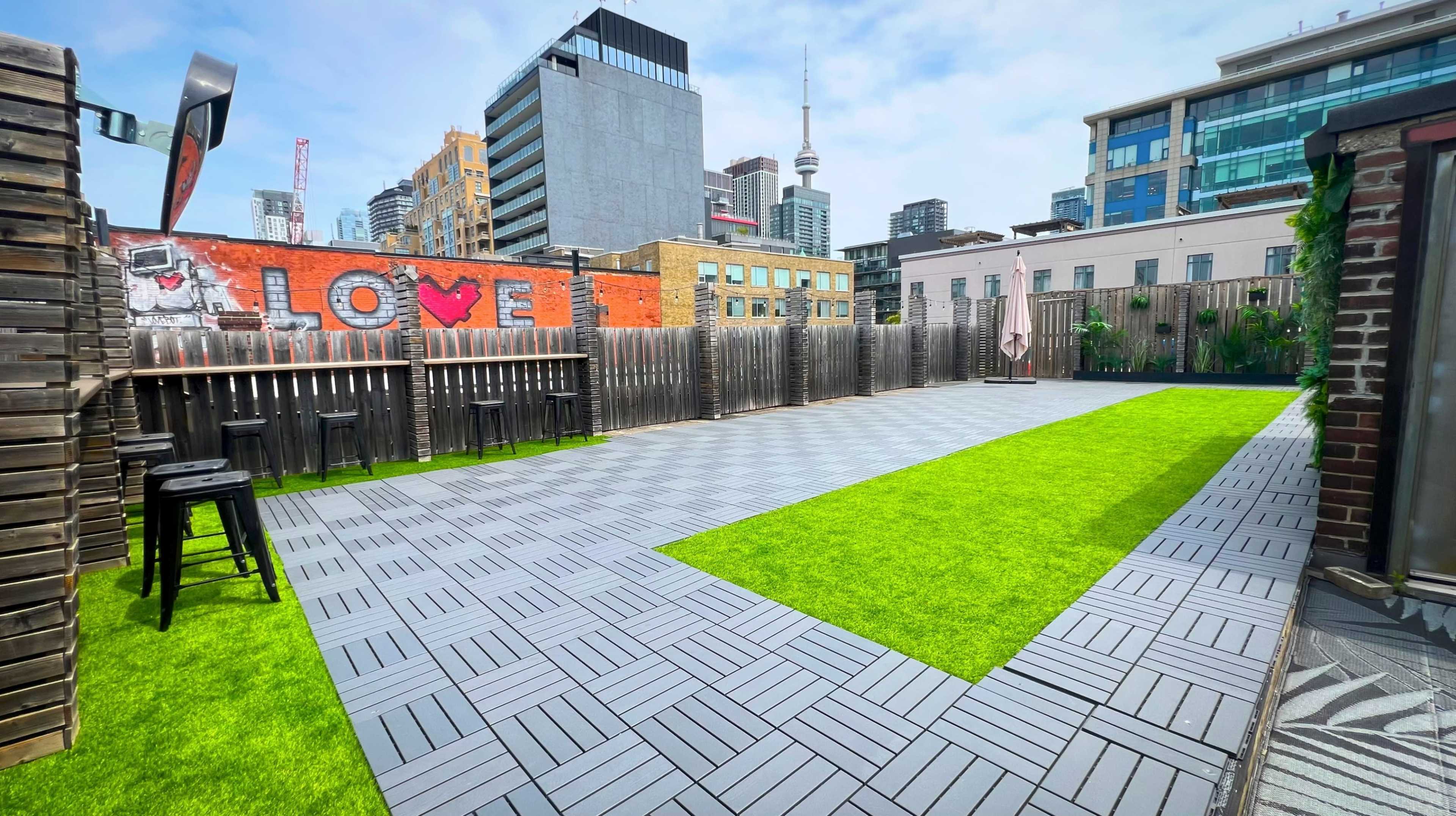 The image shows a rooftop space featuring a green artificial grass area and gray paving stones, surrounded by city buildings with a mural that has the word "LOVE."