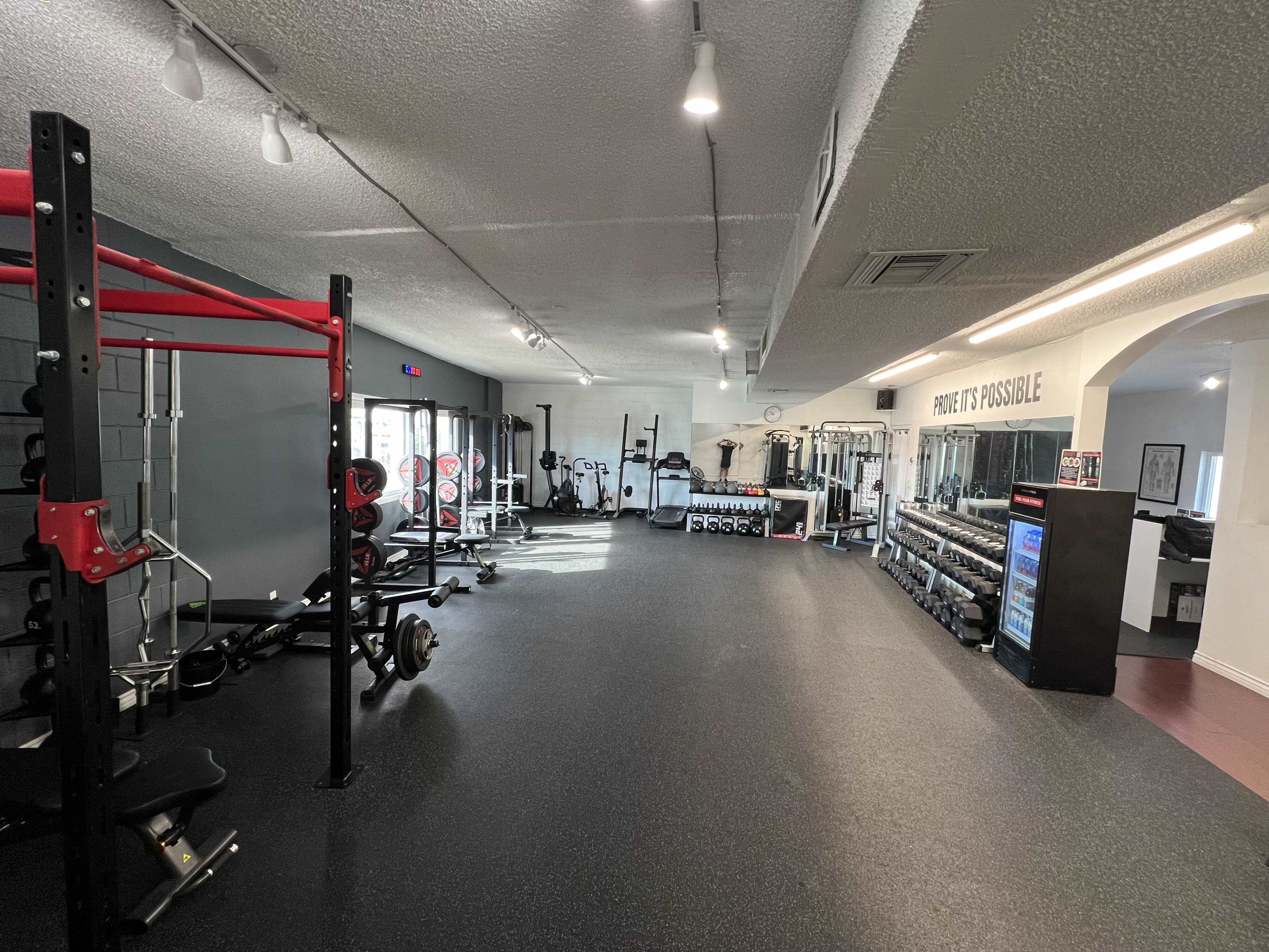 The image shows a well-equipped gym with various weightlifting and exercise machines, including a squat rack, dumbbells, and cardio equipment.