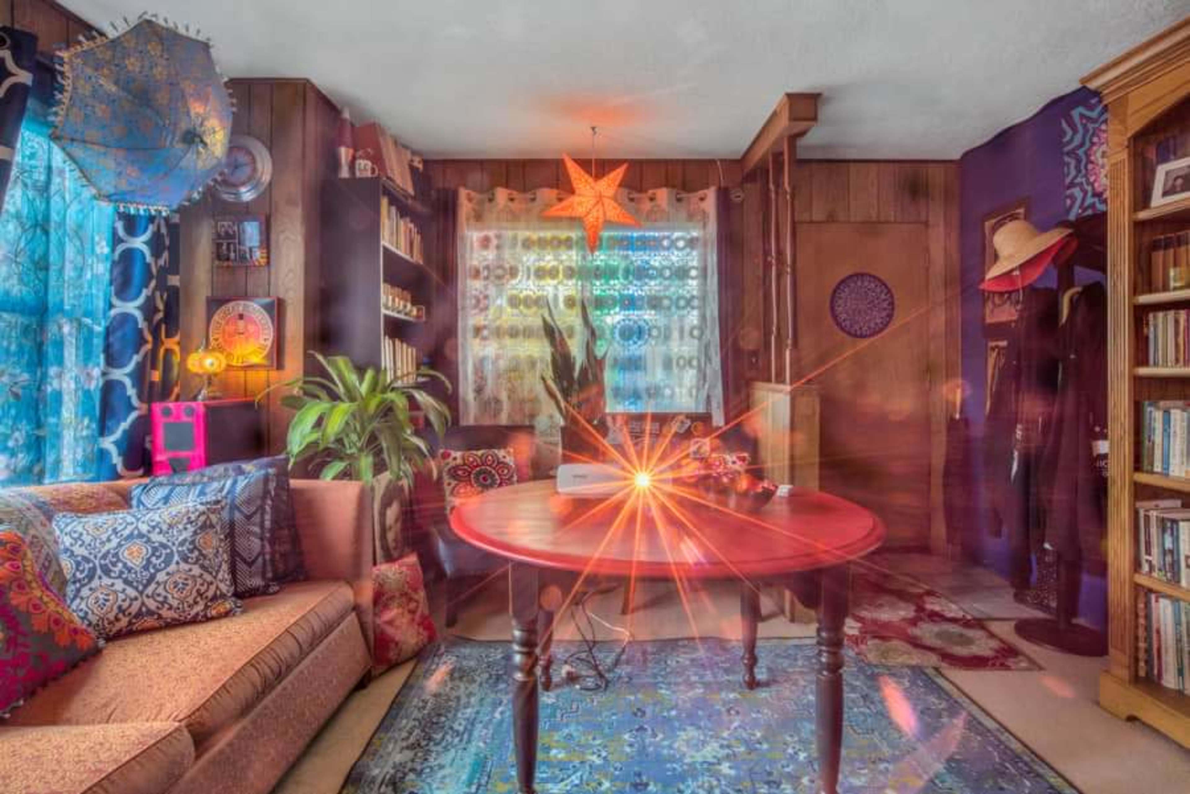 A cozy living room with a circular wooden table, a large couch, colorful patterned textiles, and shelves filled with books.
