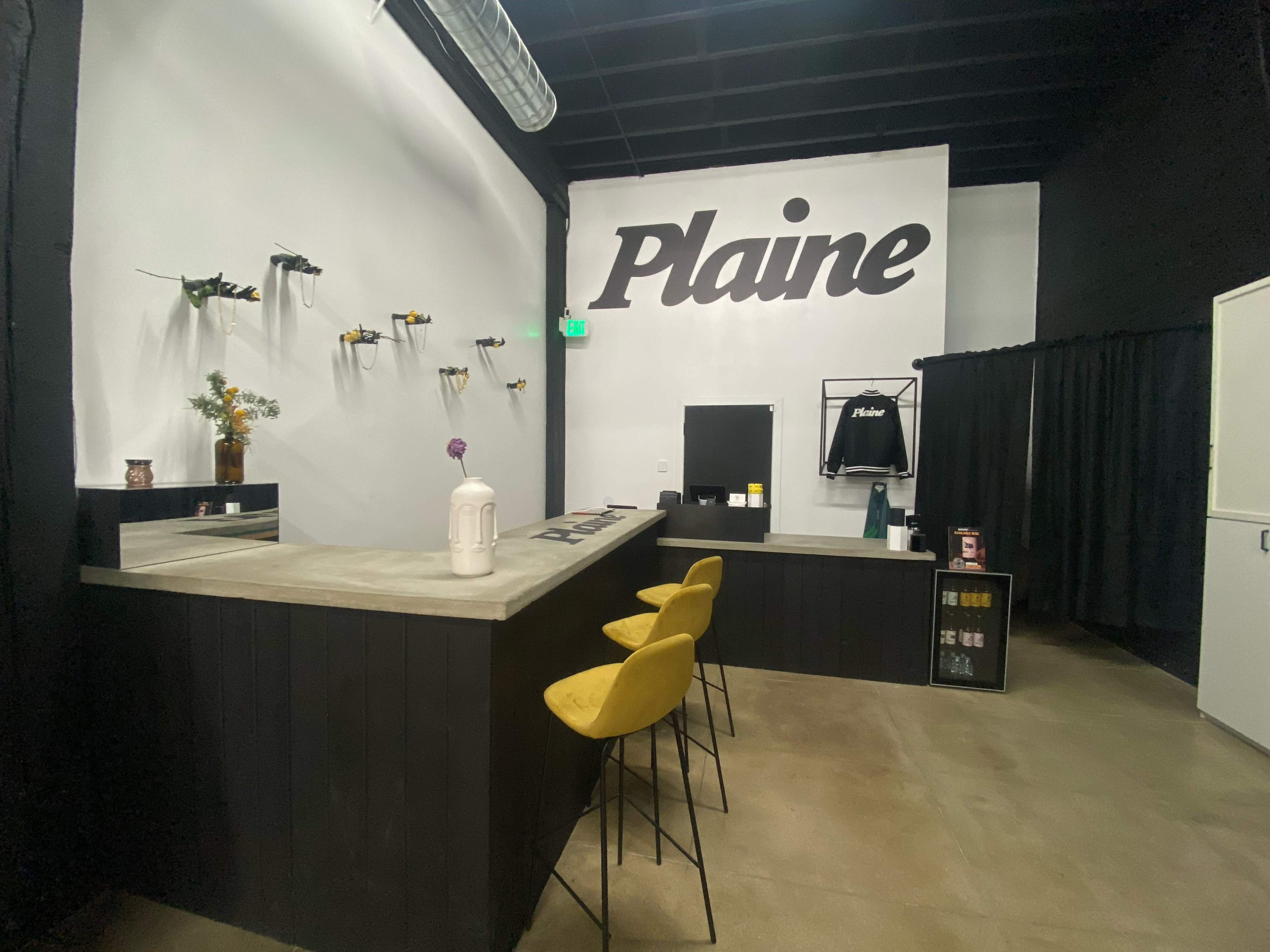 The image shows a minimalist interior of a shop with a bar-style counter, yellow stools, a large "Plaine" sign on the wall, and decorative plants on shelves.