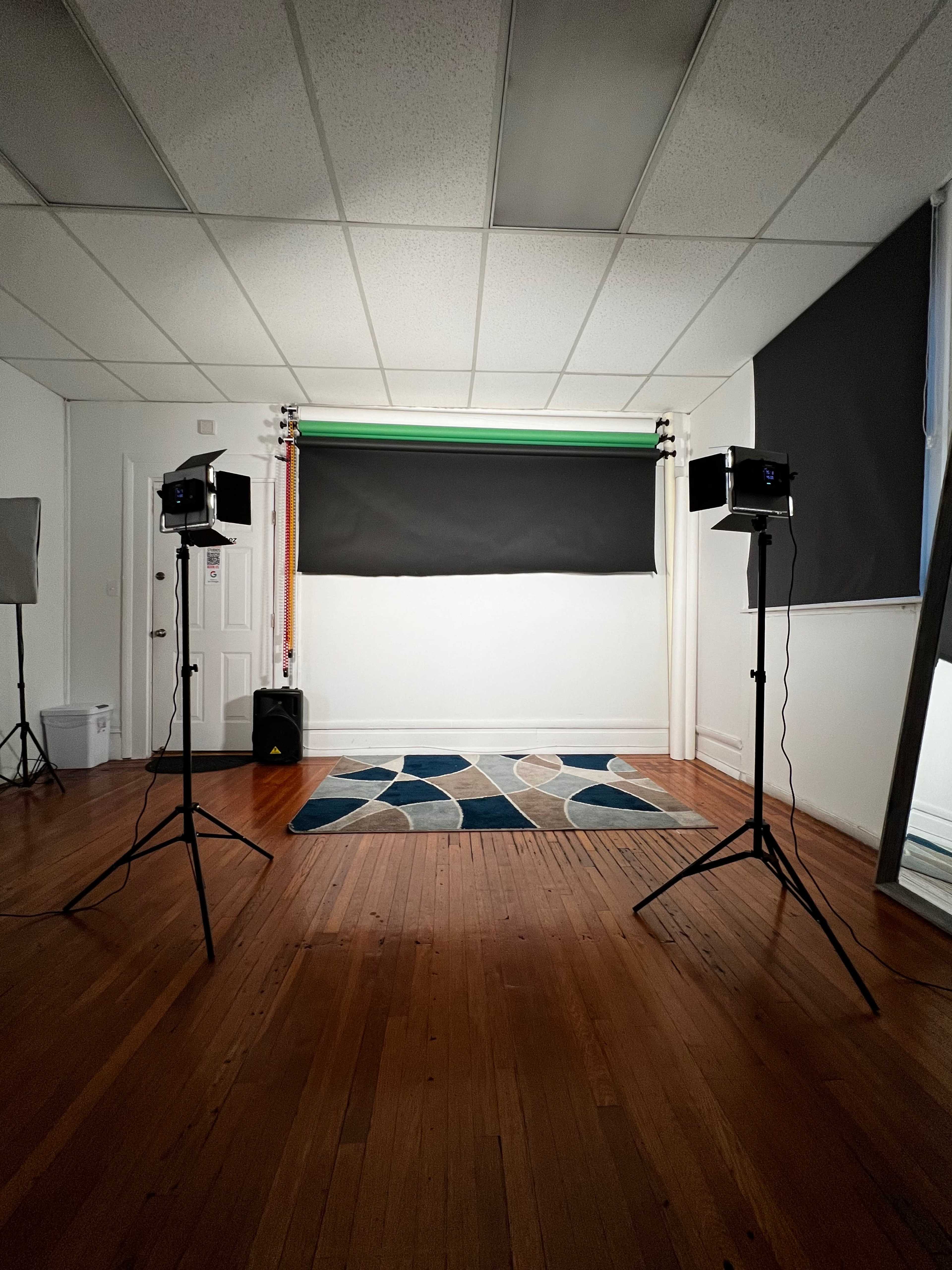 A well-lit photography studio features two lighting stands and a patterned rug on a wooden floor, with a backdrop setup against a plain wall.