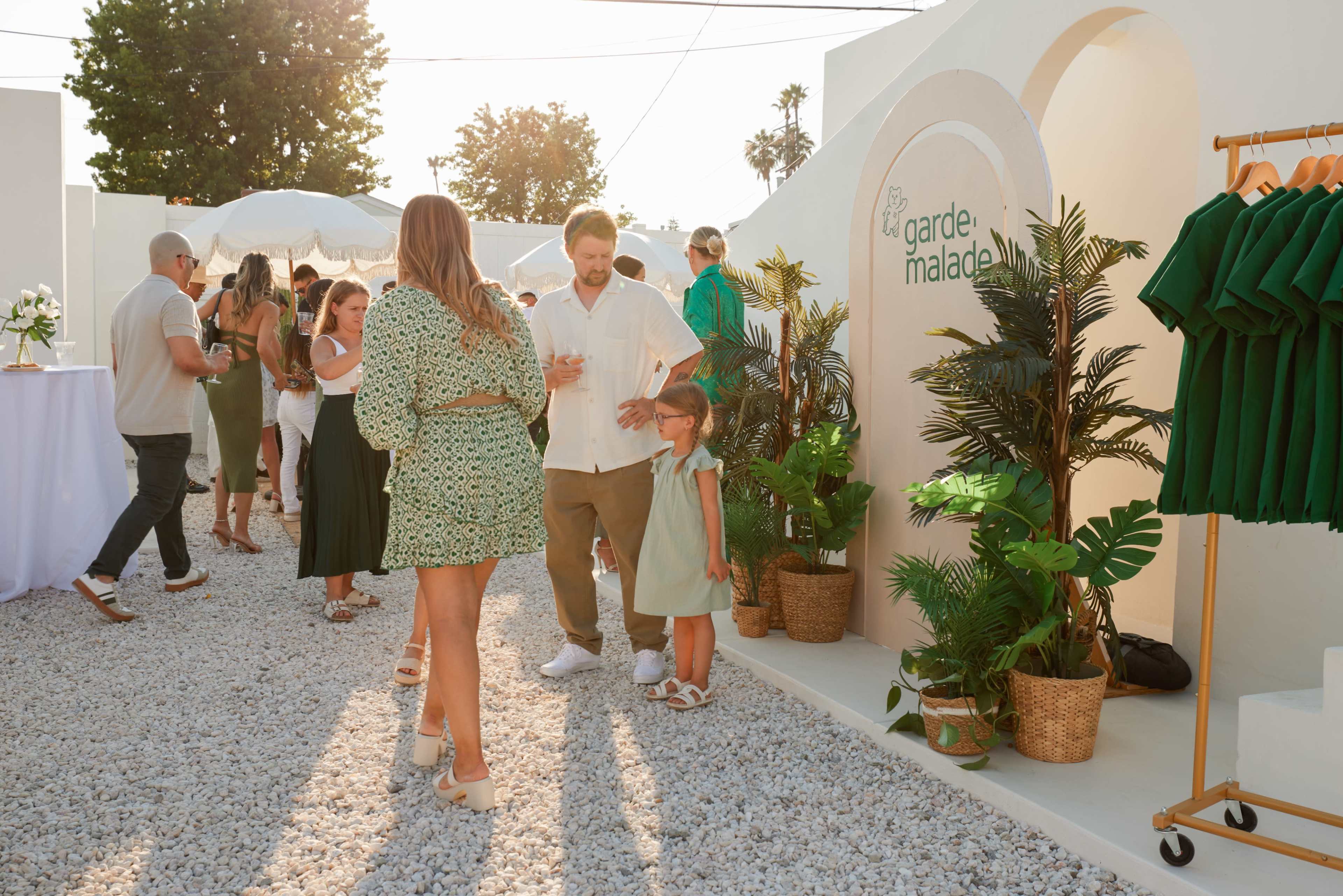 A group of people mingles at an outdoor event featuring white decor, greenery, and a sign for "la garde malade."