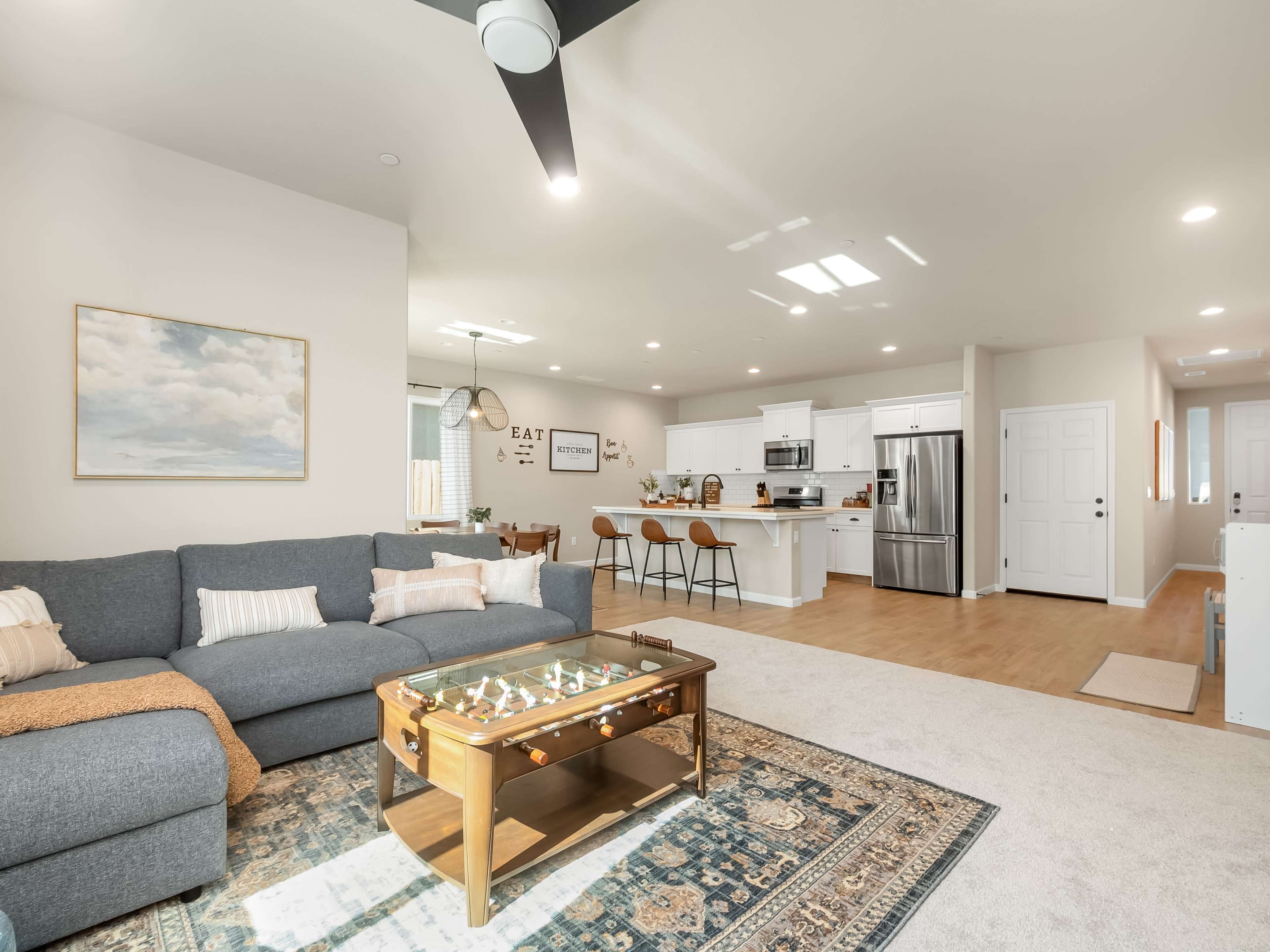 A modern living room features a gray sectional sofa, a glass-top coffee table, and an open kitchen area with bar seating.