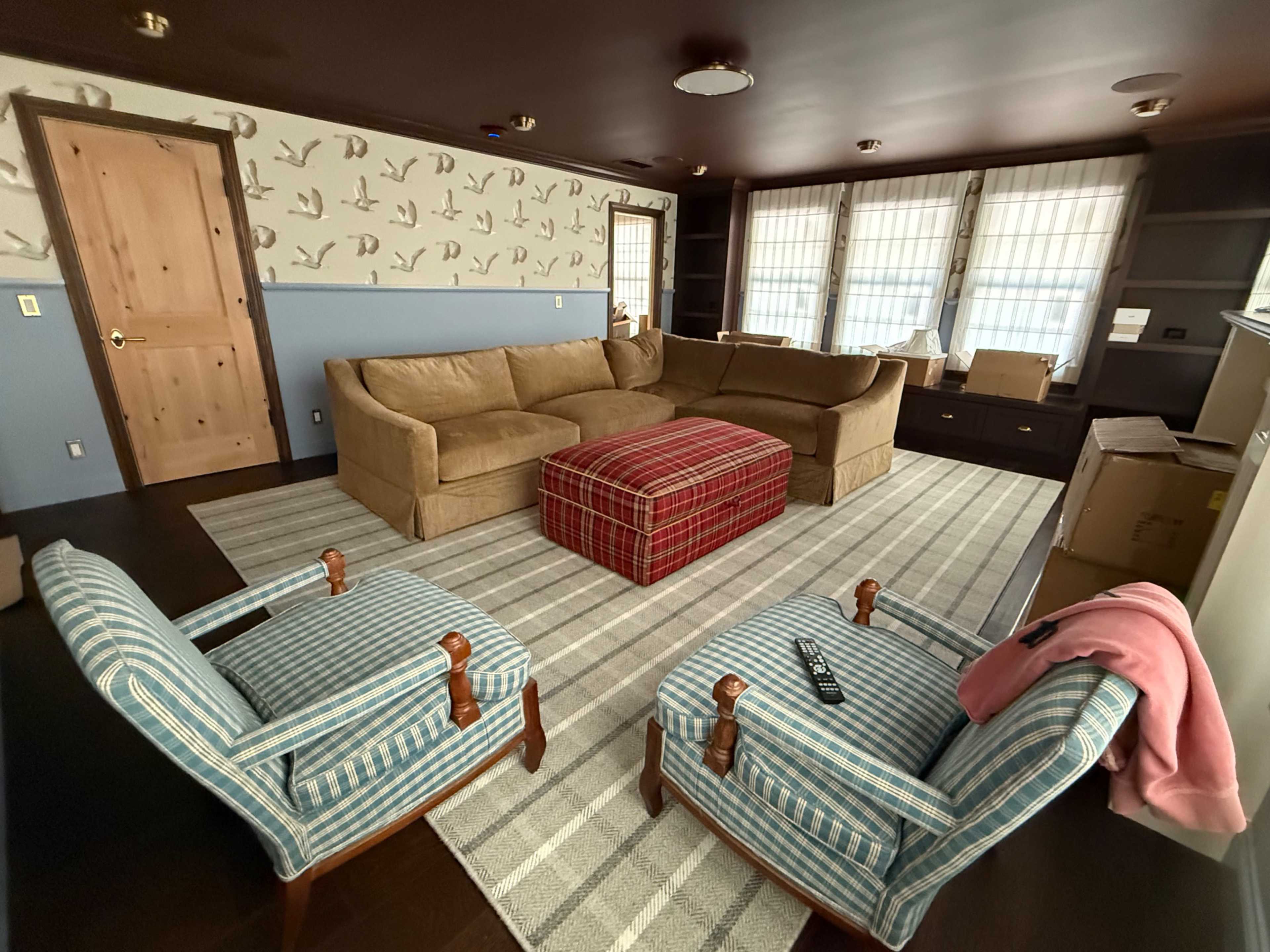 The room features a brown sectional sofa, two blue-and-white striped chairs, a plaid coffee table, and boxes against the walls.