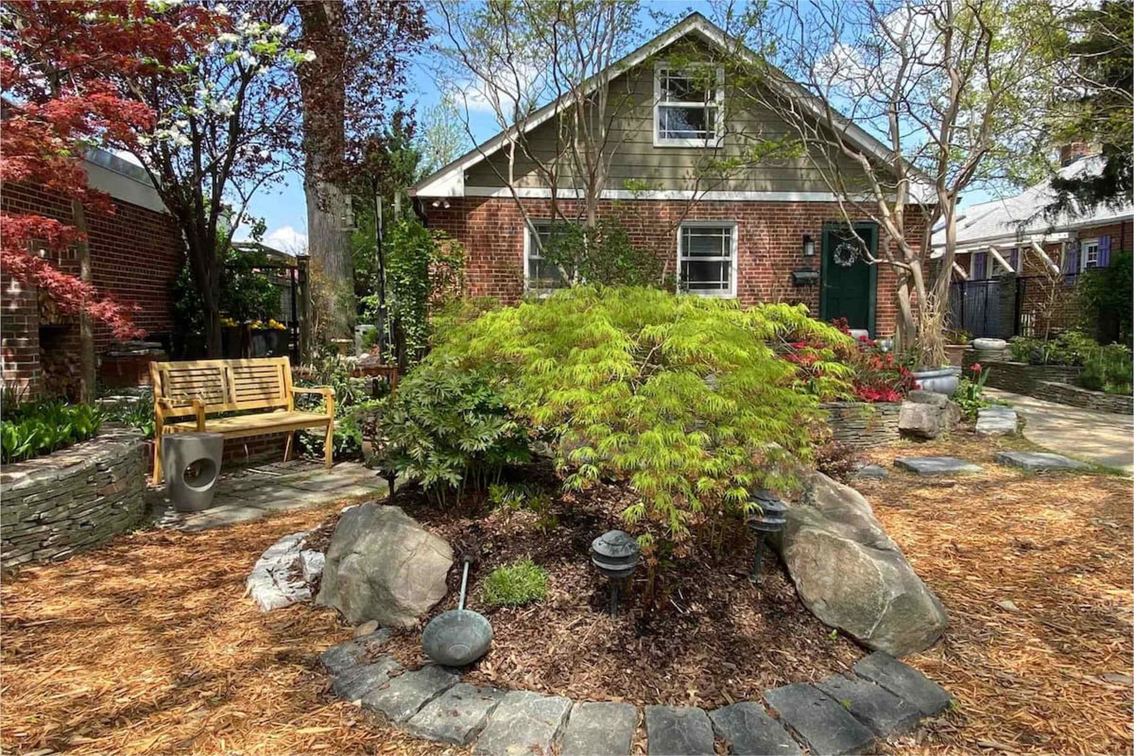 A brick house with a green door is surrounded by a landscaped garden featuring a stone pathway, plants, and a wooden bench.