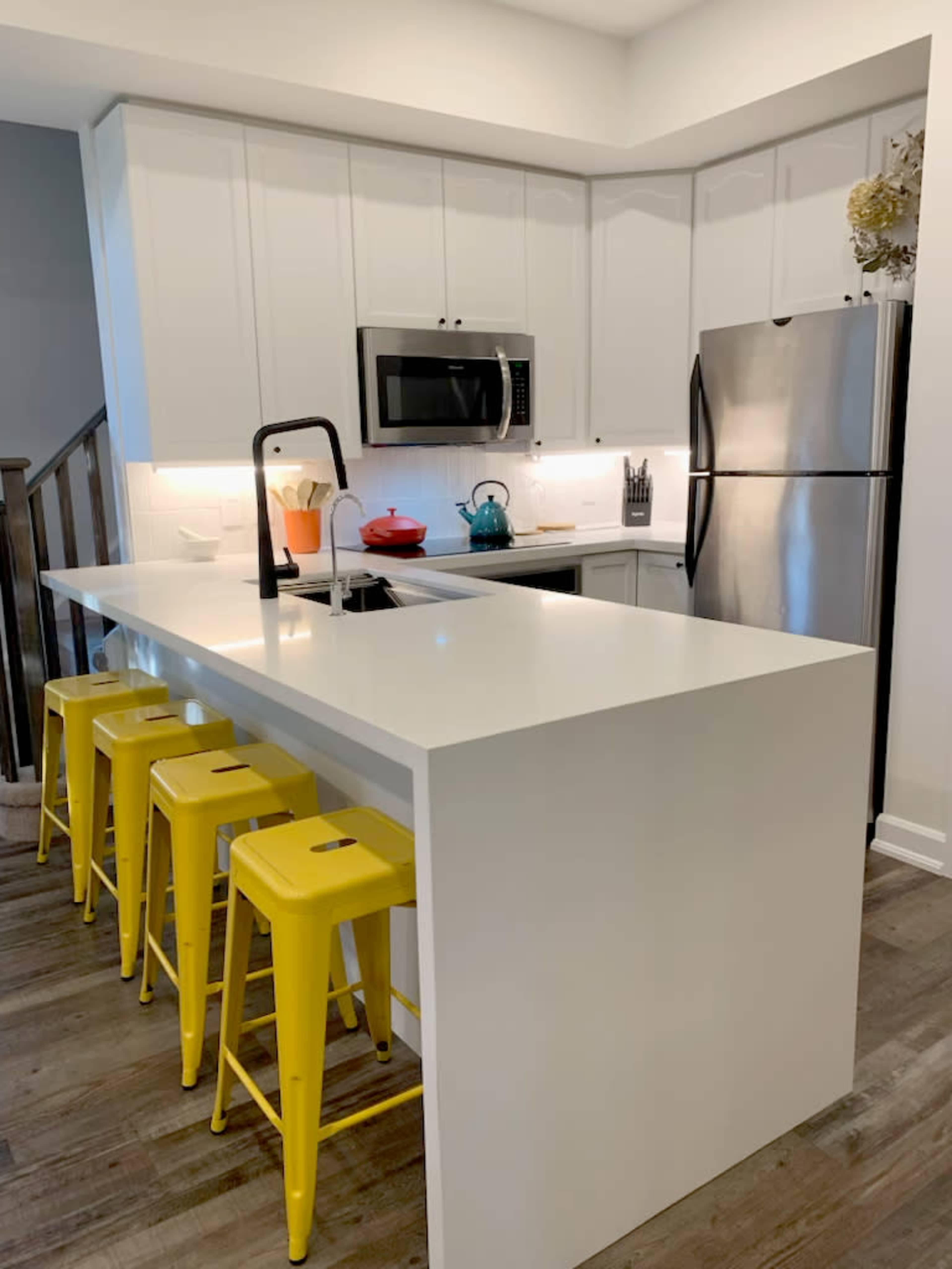 The image shows a modern kitchen with white cabinets, stainless steel appliances, and four yellow stools at a white island.