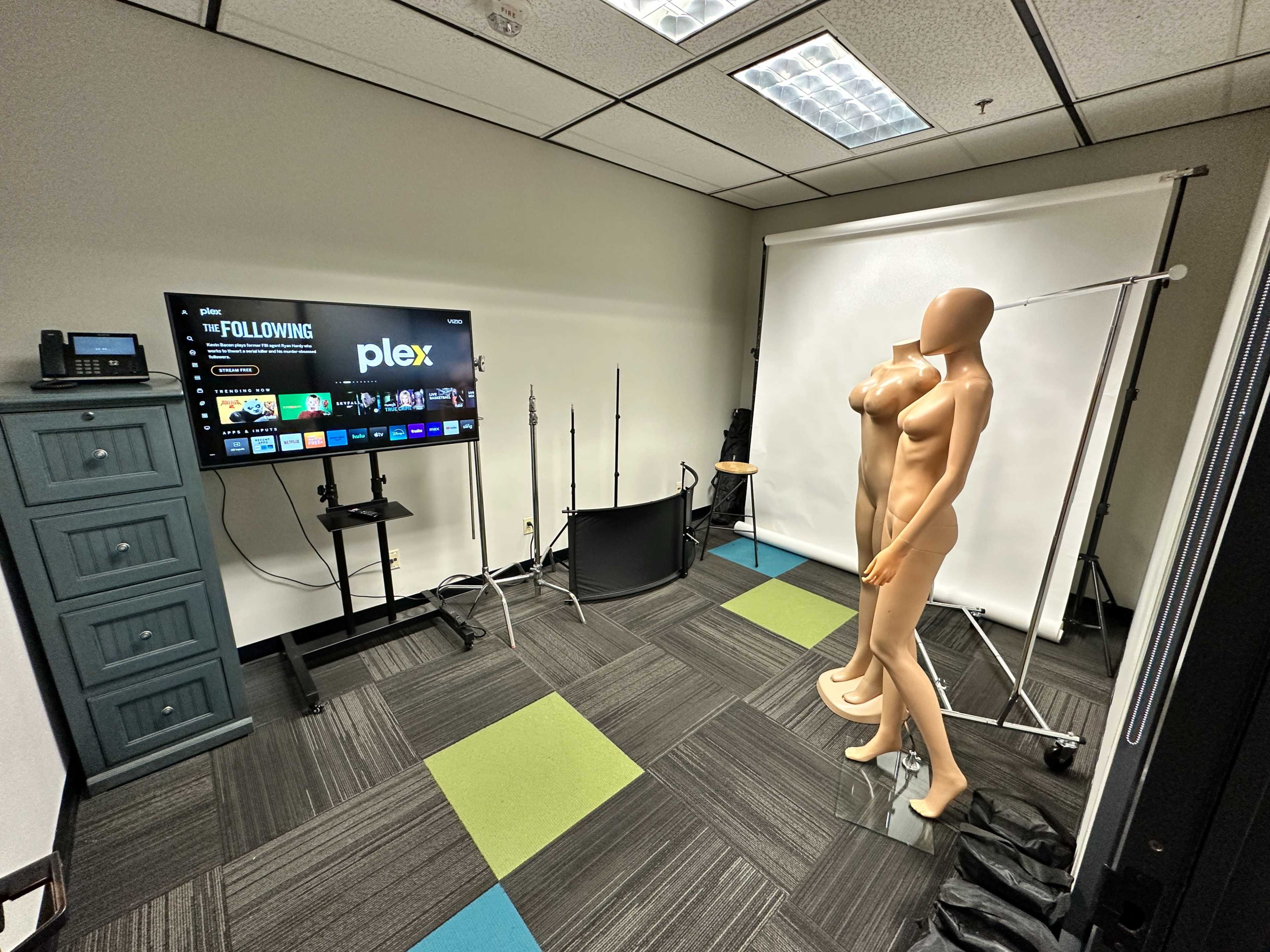 The image shows a small room with a television displaying the Plex interface, a blue dresser, a pair of mannequin torsos, and lighting equipment set up on a backdrop.