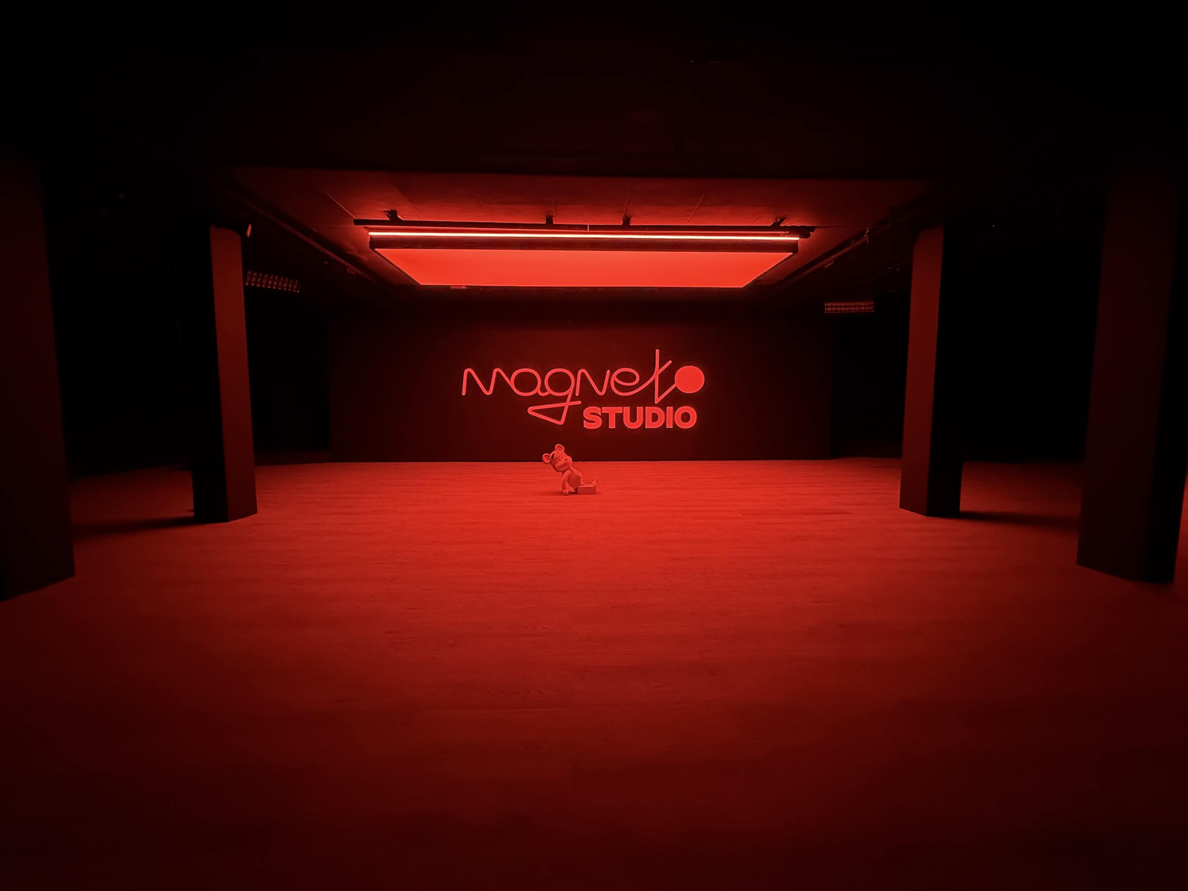 A dimly lit studio space with a red hue, featuring the words "Magneto Studio" on the wall and a lone object in the center.
