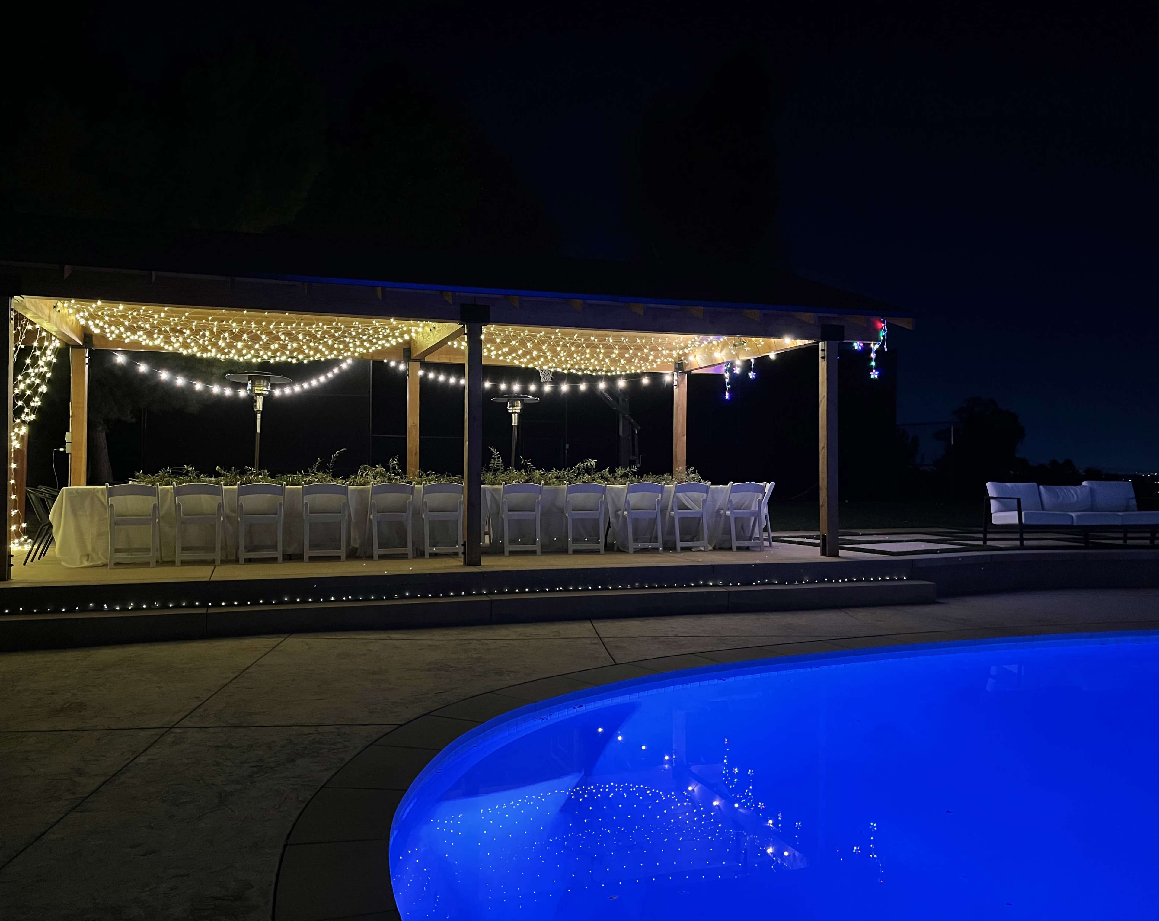 A illuminated outdoor dining area with a long table set for a gathering stands next to a brightly lit swimming pool at night.