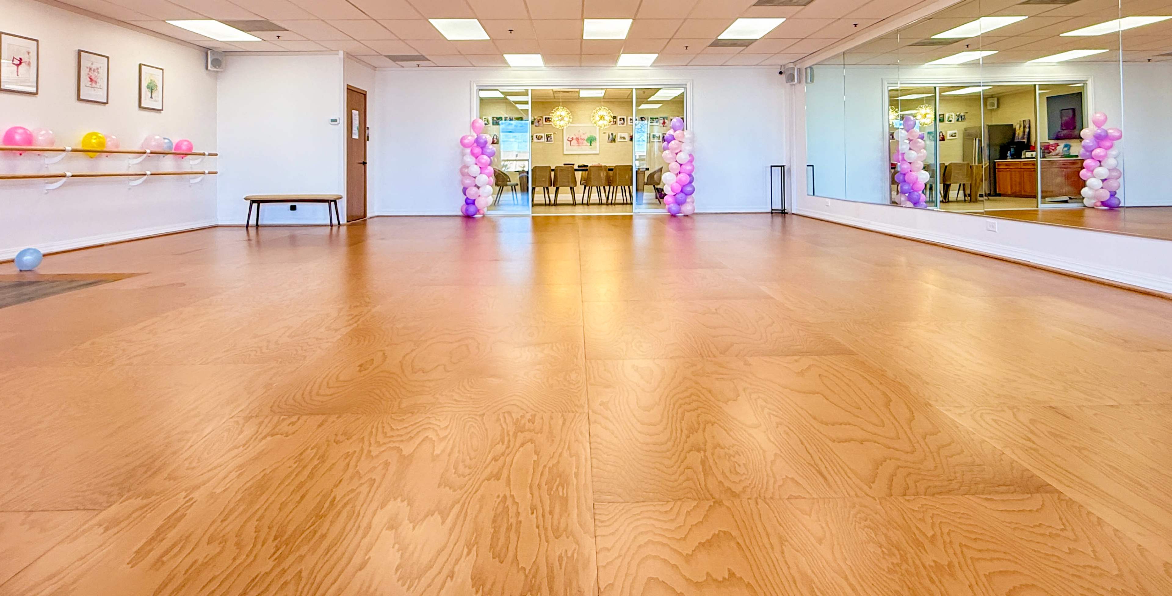 The image shows a large, empty room with wooden flooring, mirrored walls, and colorful balloon decorations at each end.