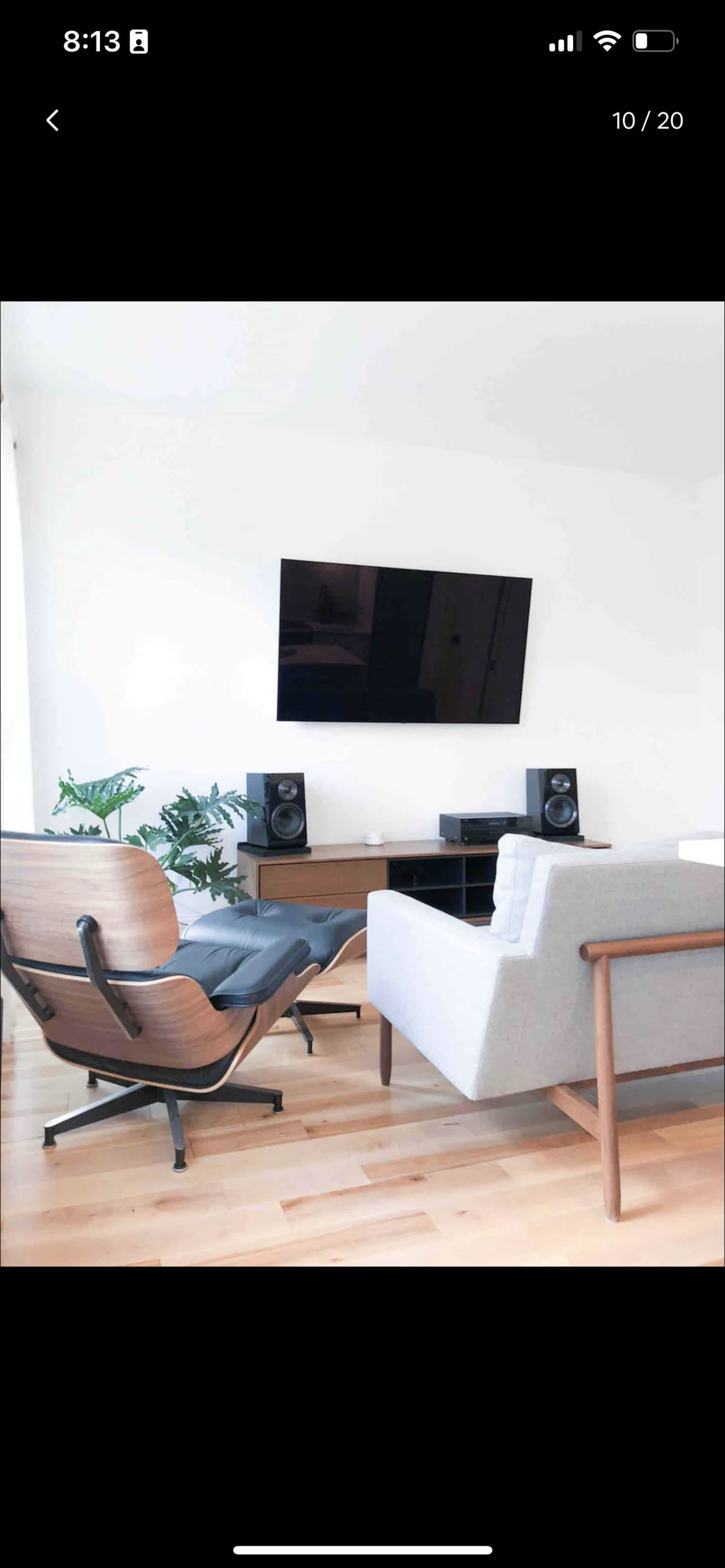 A modern living room features a gray sofa, an Eames-style chair, a television mounted on the white wall, and speakers on a wooden media console.