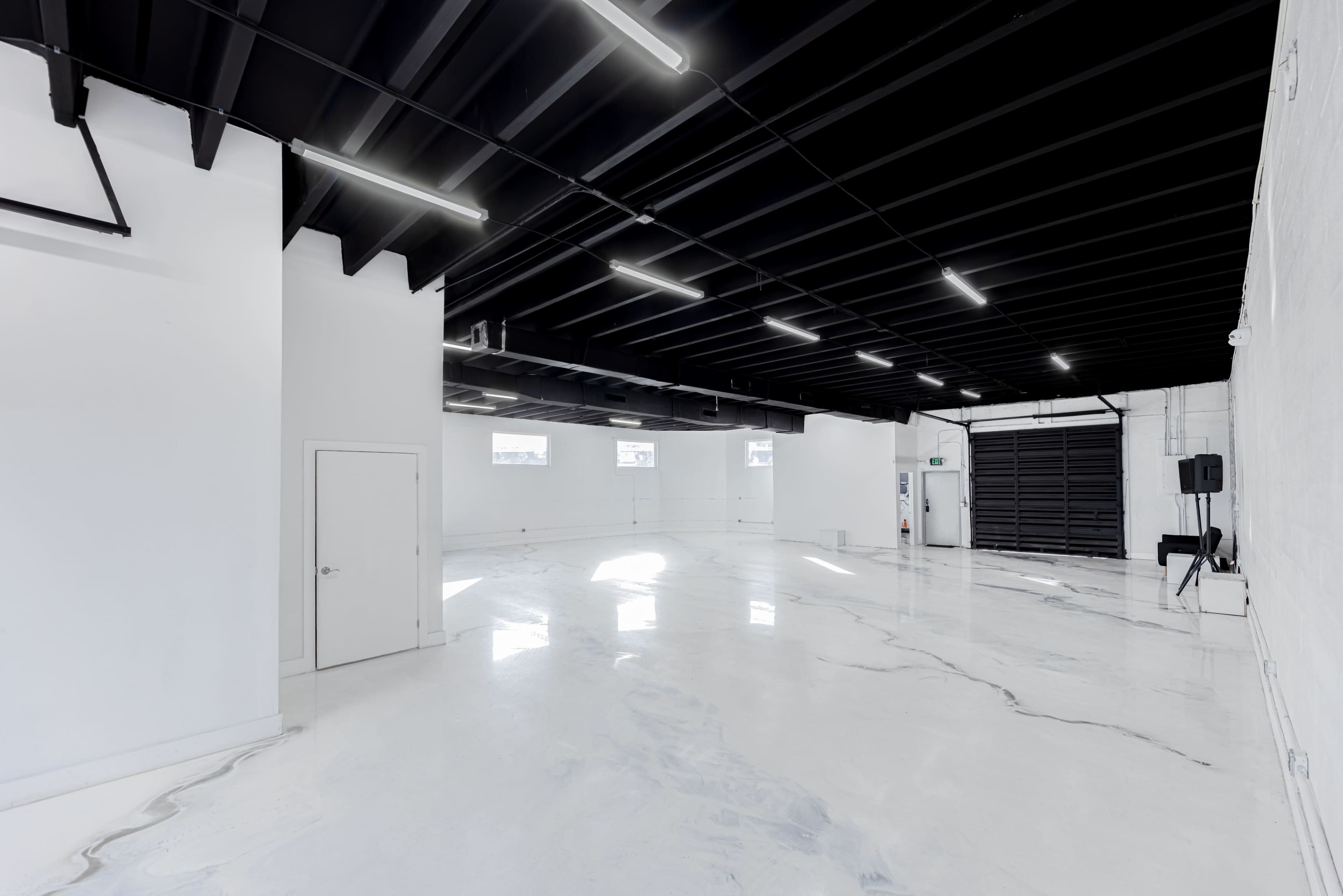The image shows a spacious, minimally furnished room with a polished white floor and black ceiling, featuring large windows and a roll-up door.