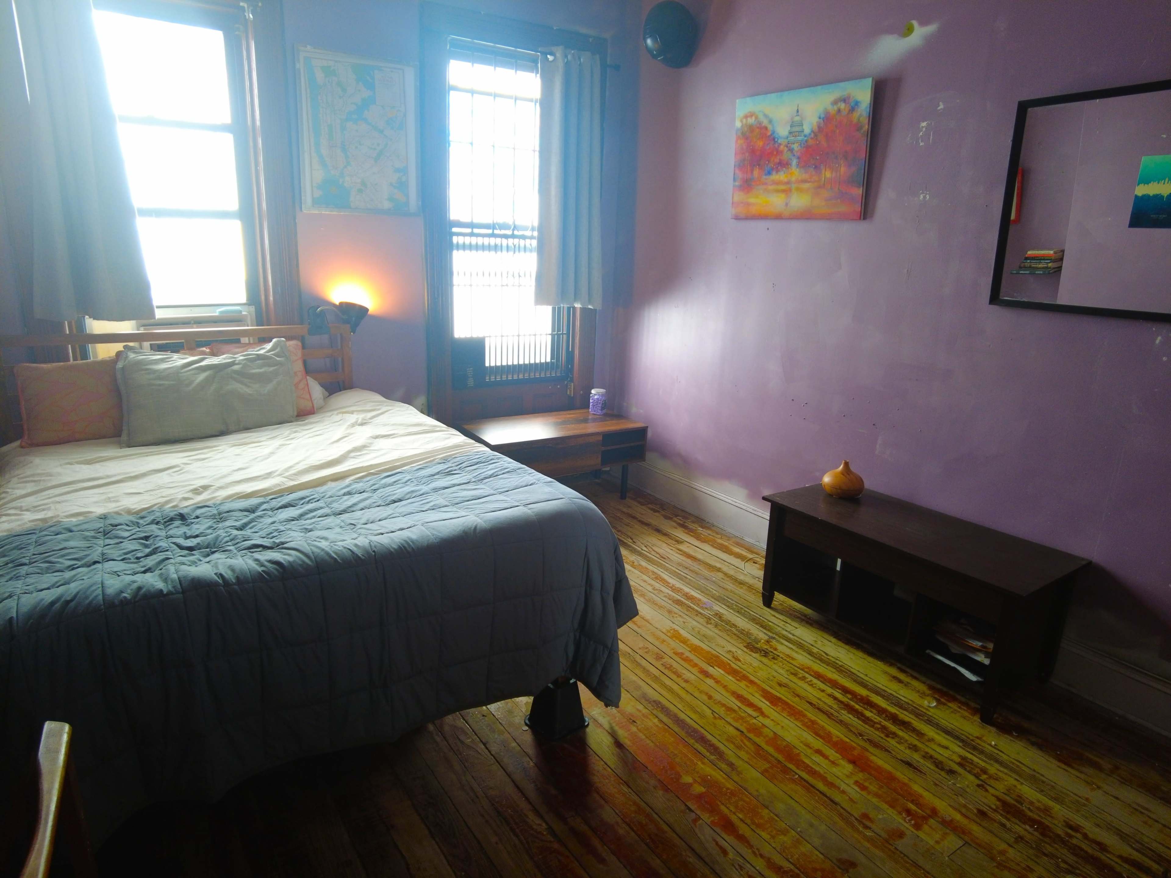 A bedroom features a bed with gray bedding, wooden floors, a nightstand, and a small television stand against a lavender wall.