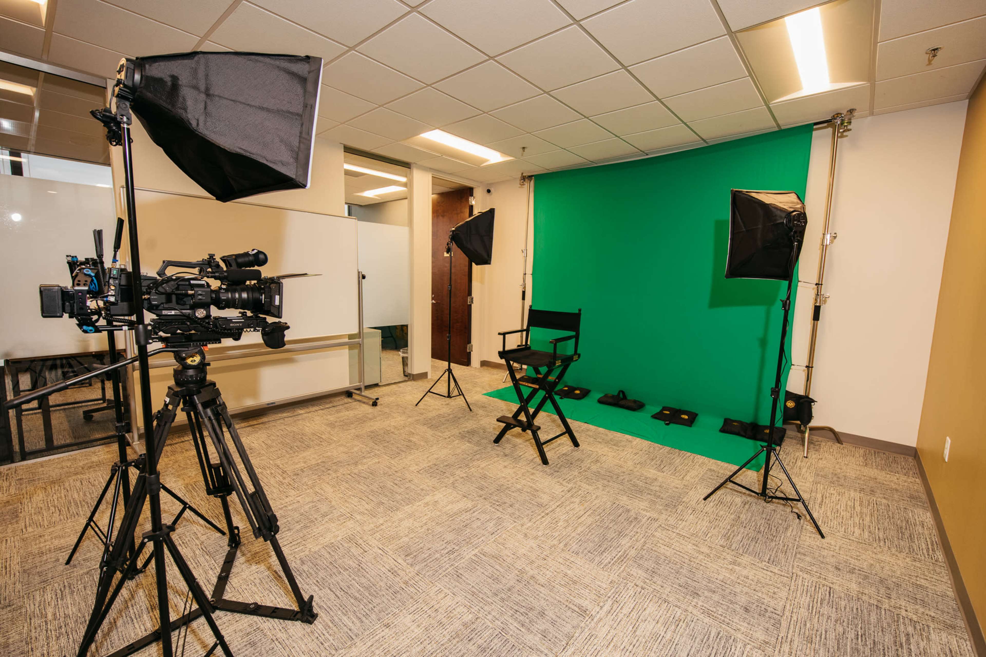 A small studio setup featuring a green screen, two lights, a video camera on a tripod, and a director's chair.