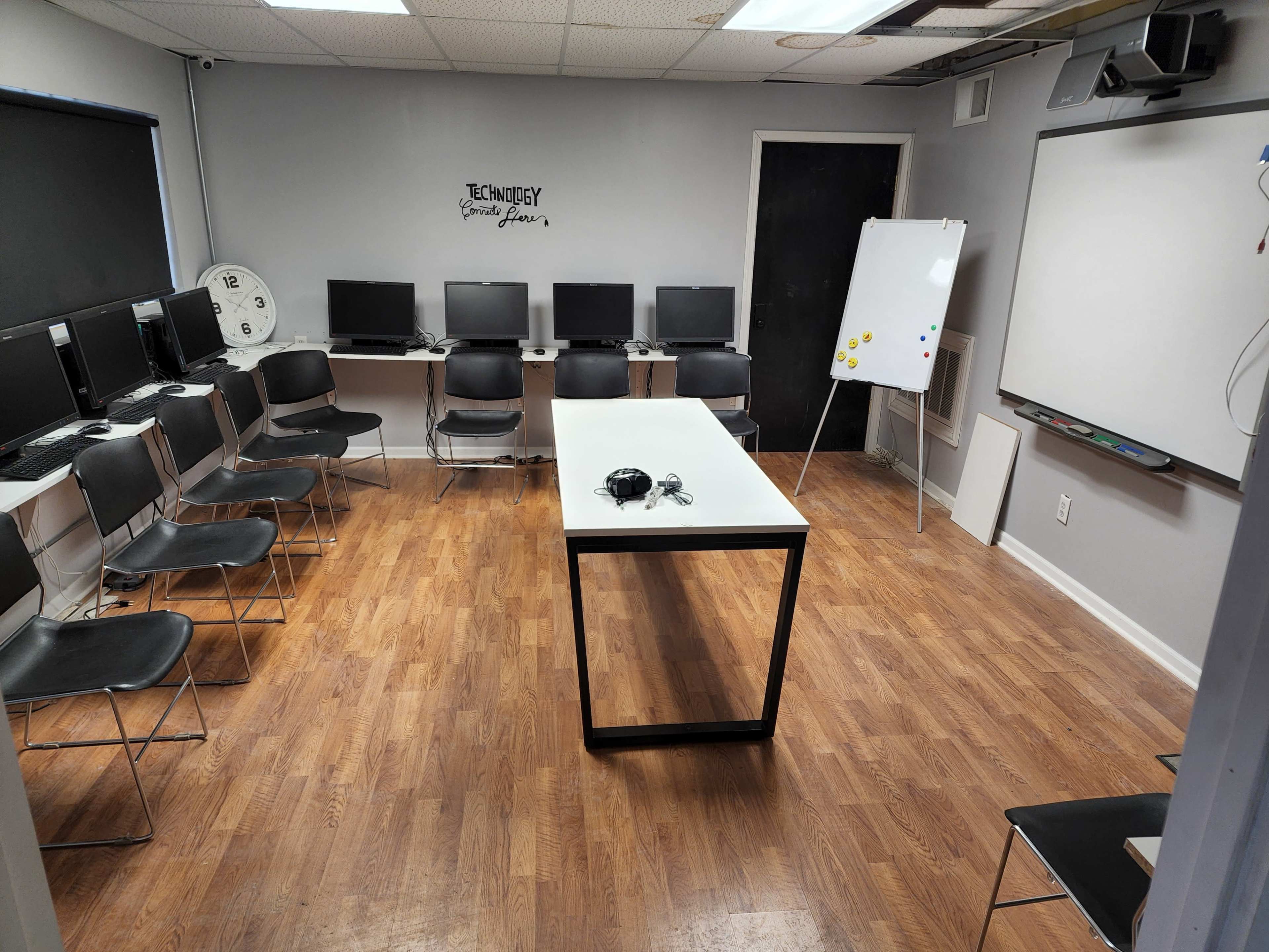 A computer lab with several desktop computers arranged at tables, a large whiteboard, and a central table surrounded by chairs.