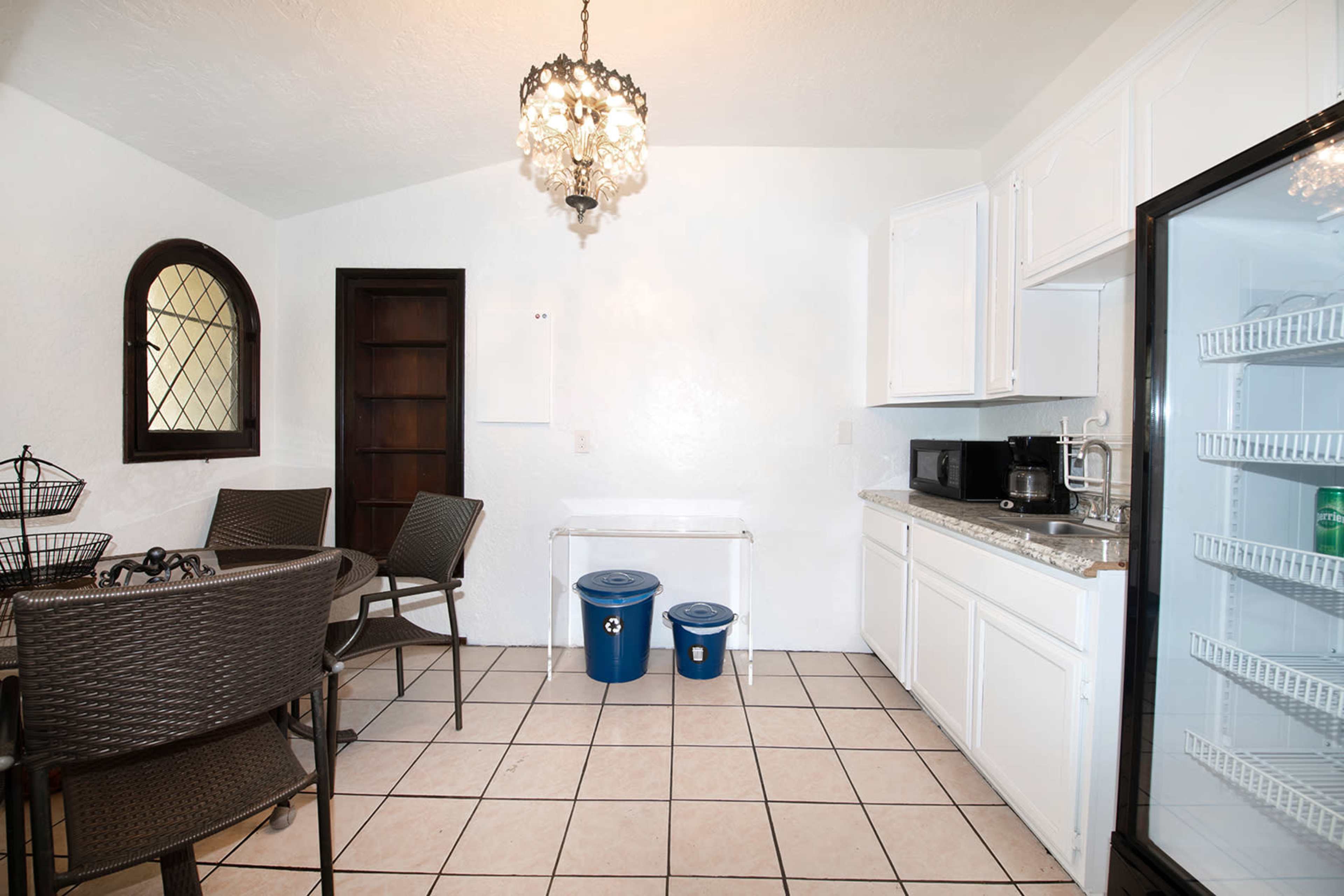 A small kitchen area with white cabinets, a refrigerator, a microwave, a table with chairs, and two blue bins on the floor.
