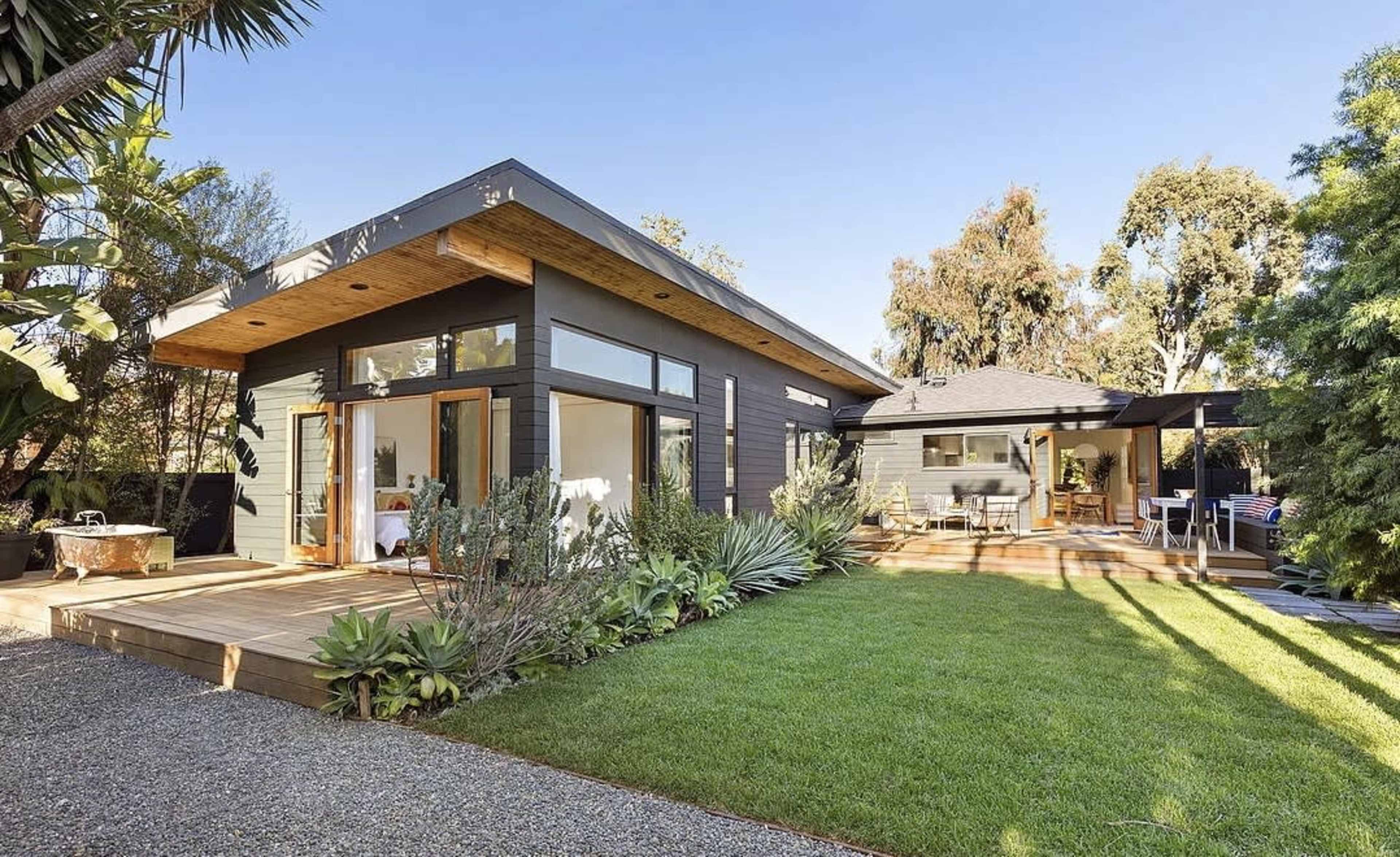 A modern single-story house features large windows, a wooden deck, and a landscaped yard with various plants.