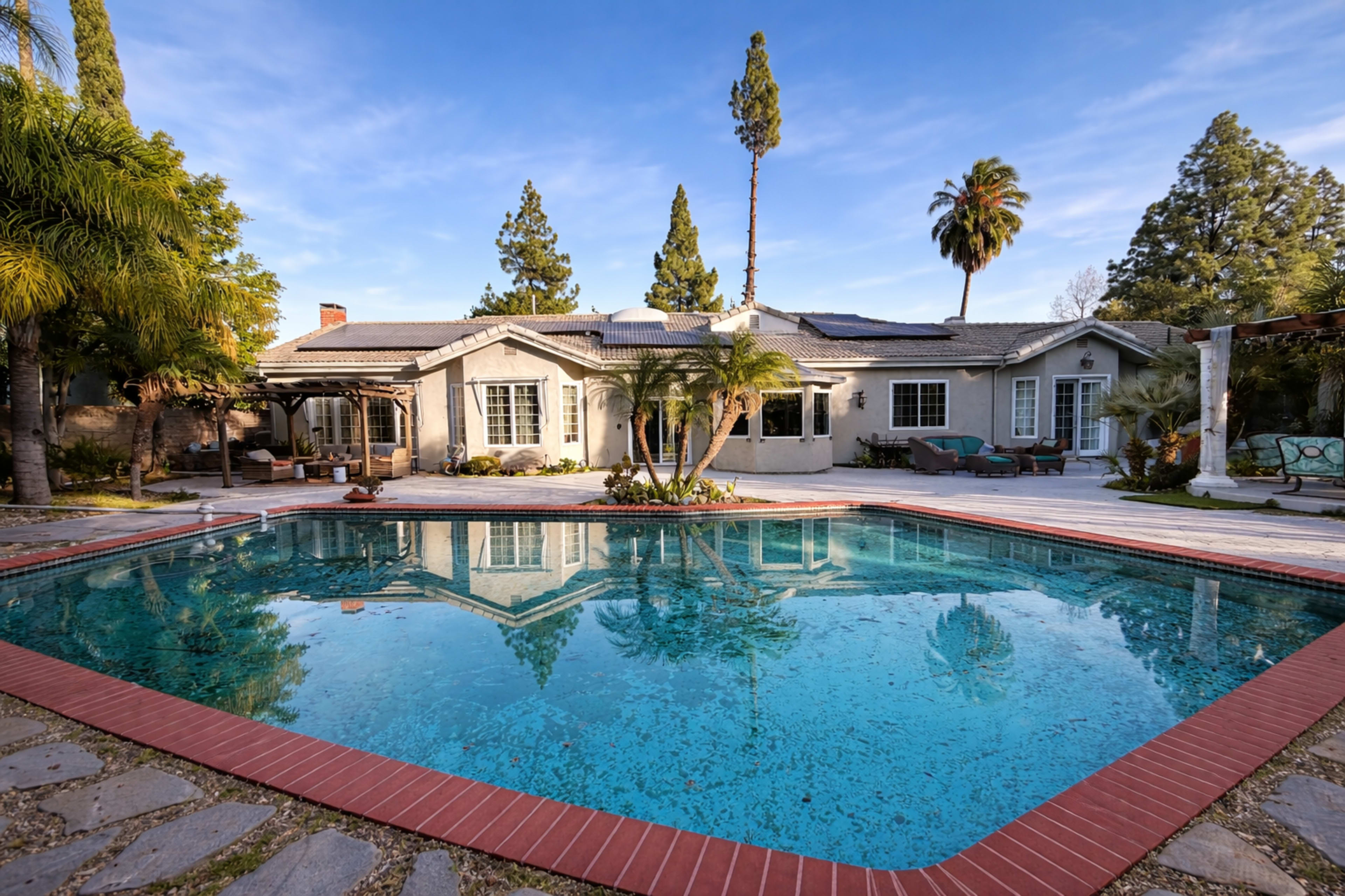 The image shows a spacious outdoor area featuring a pool in front of a single-story house surrounded by trees and patio furniture.