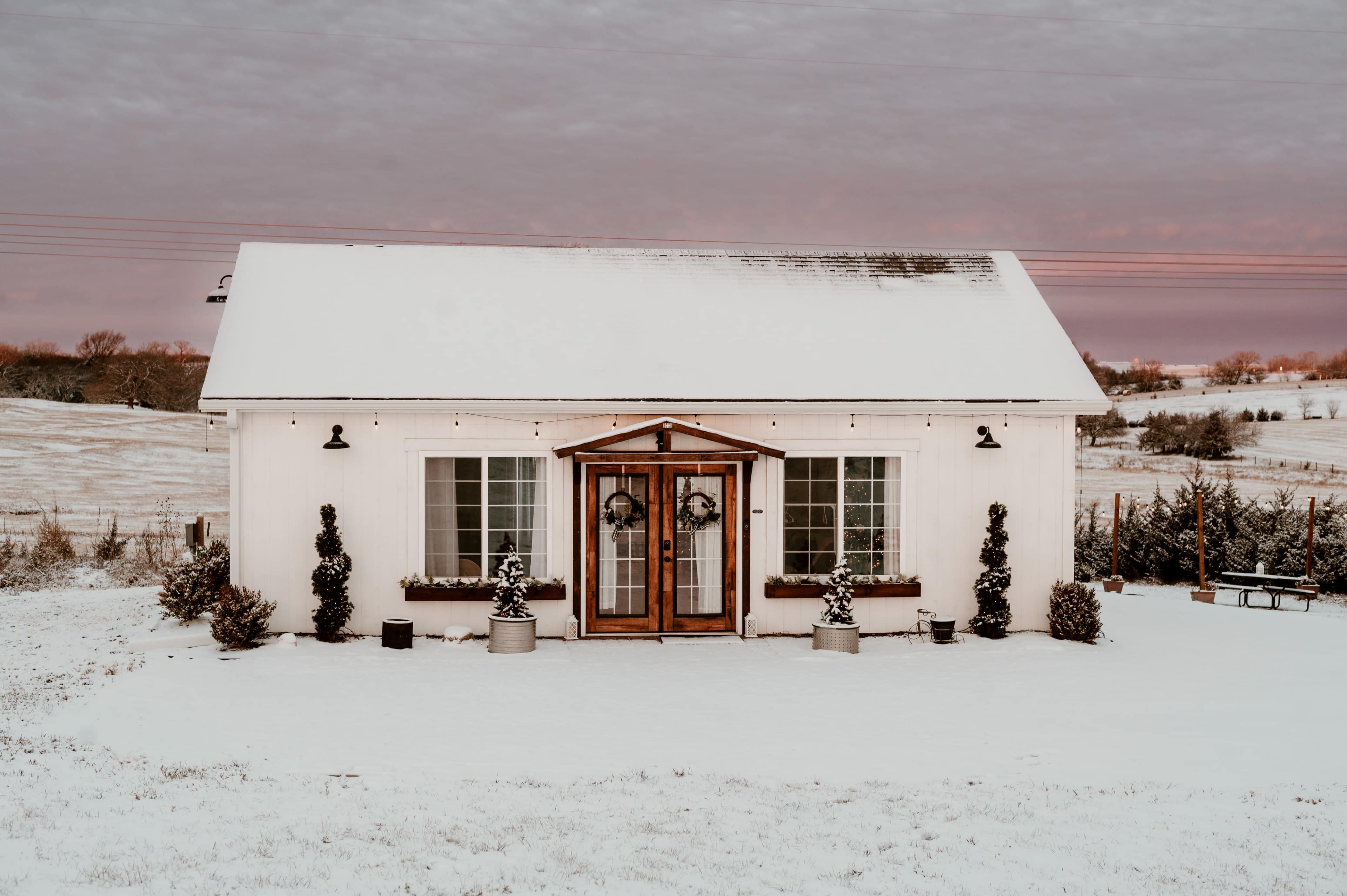 A snow-covered house with wooden double doors and potted plants stands in a winter landscape.