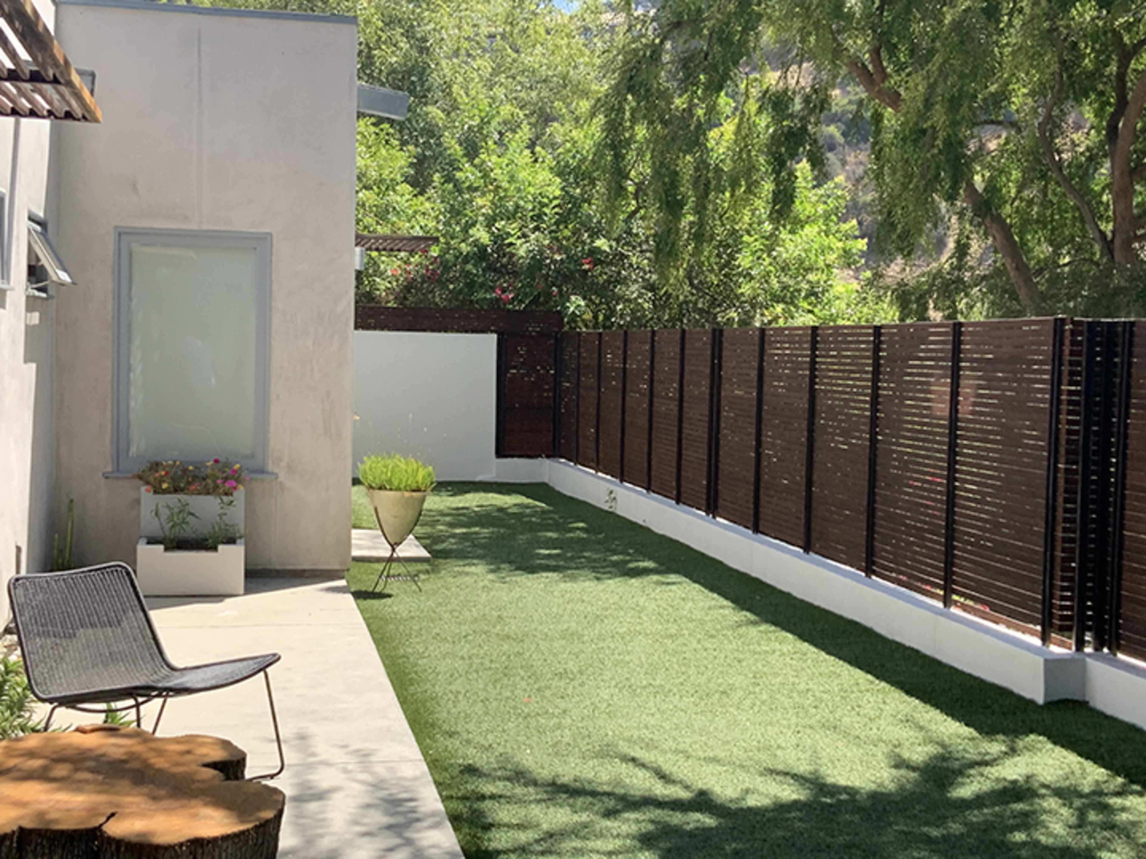 The image shows a modern backyard with artificial grass, a wooden privacy fence, a garden area with a planter, and a chair positioned on a concrete patio.