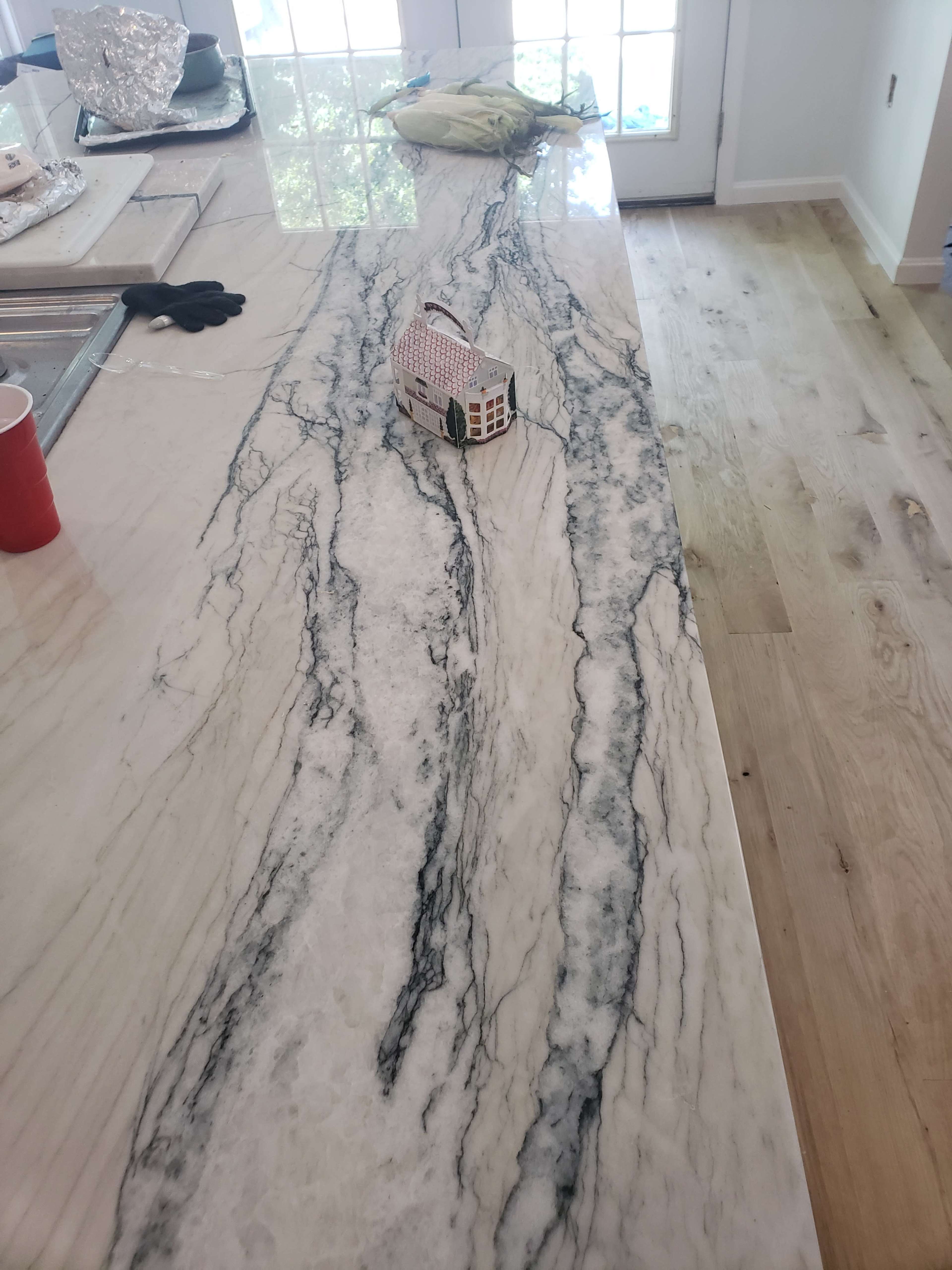 A marble countertop with a swirling black and white pattern displays a small toy house on its surface, alongside a red cup and a piece of green produce.