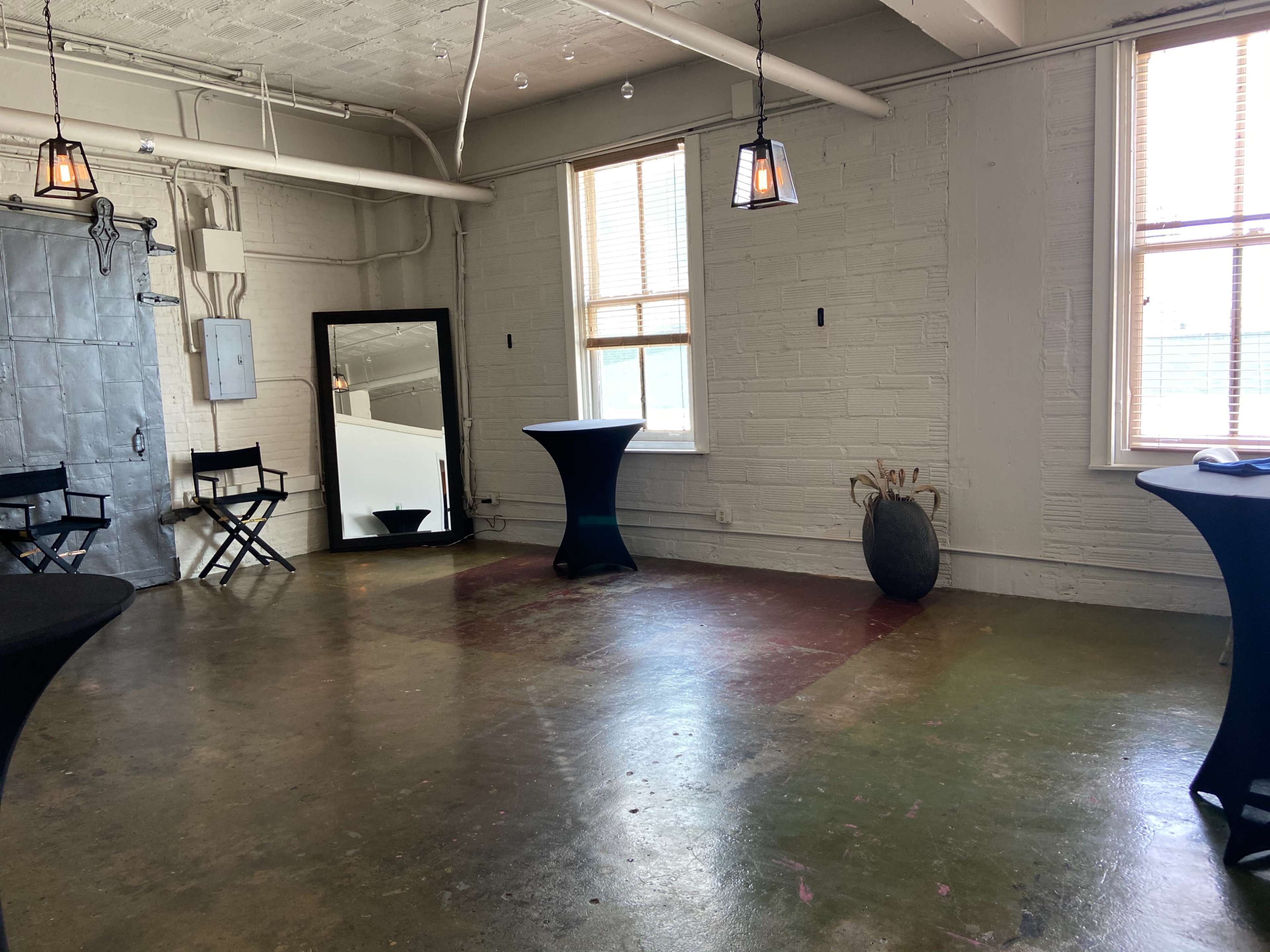 A spacious room with two windows, a large mirror, tall black tables, director's chairs, and a decorative vase, all against a textured white wall and a polished concrete floor.
