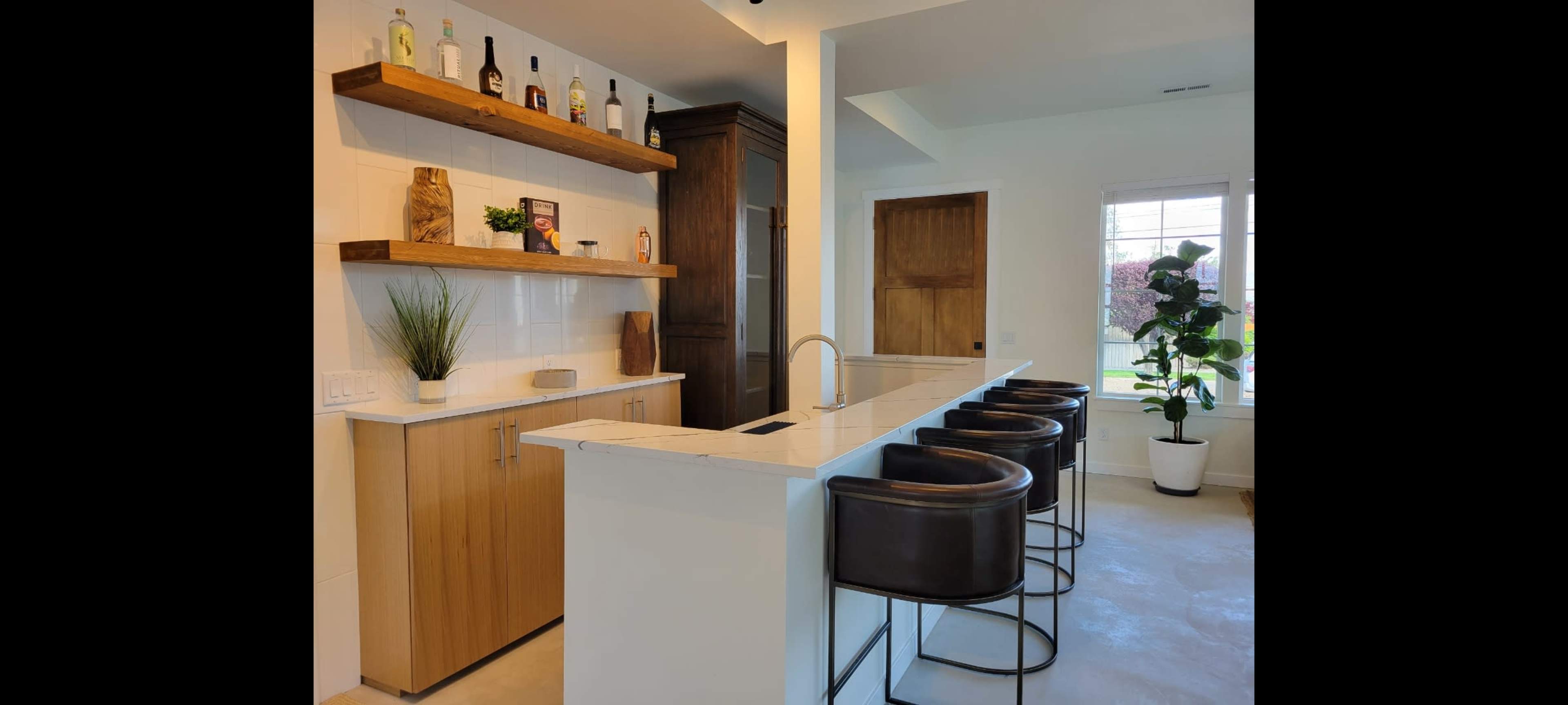 A modern kitchen bar area with a white countertop, brown bar stools, wooden shelves displaying bottles, and a potted plant near a window.