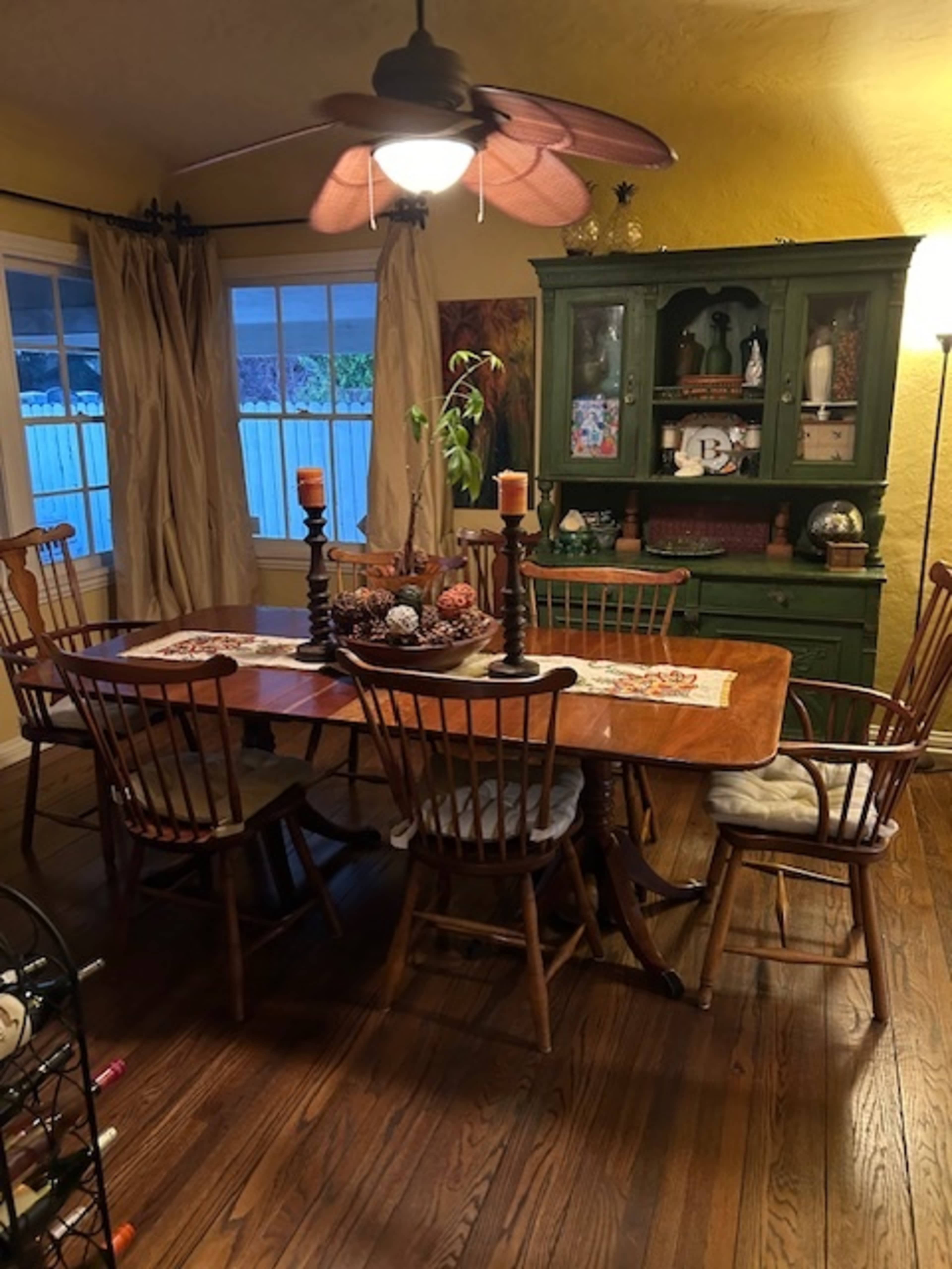 A wooden dining table is set in a room with a green china cabinet, seven chairs, and a ceiling fan, complemented by large windows and yellow walls.