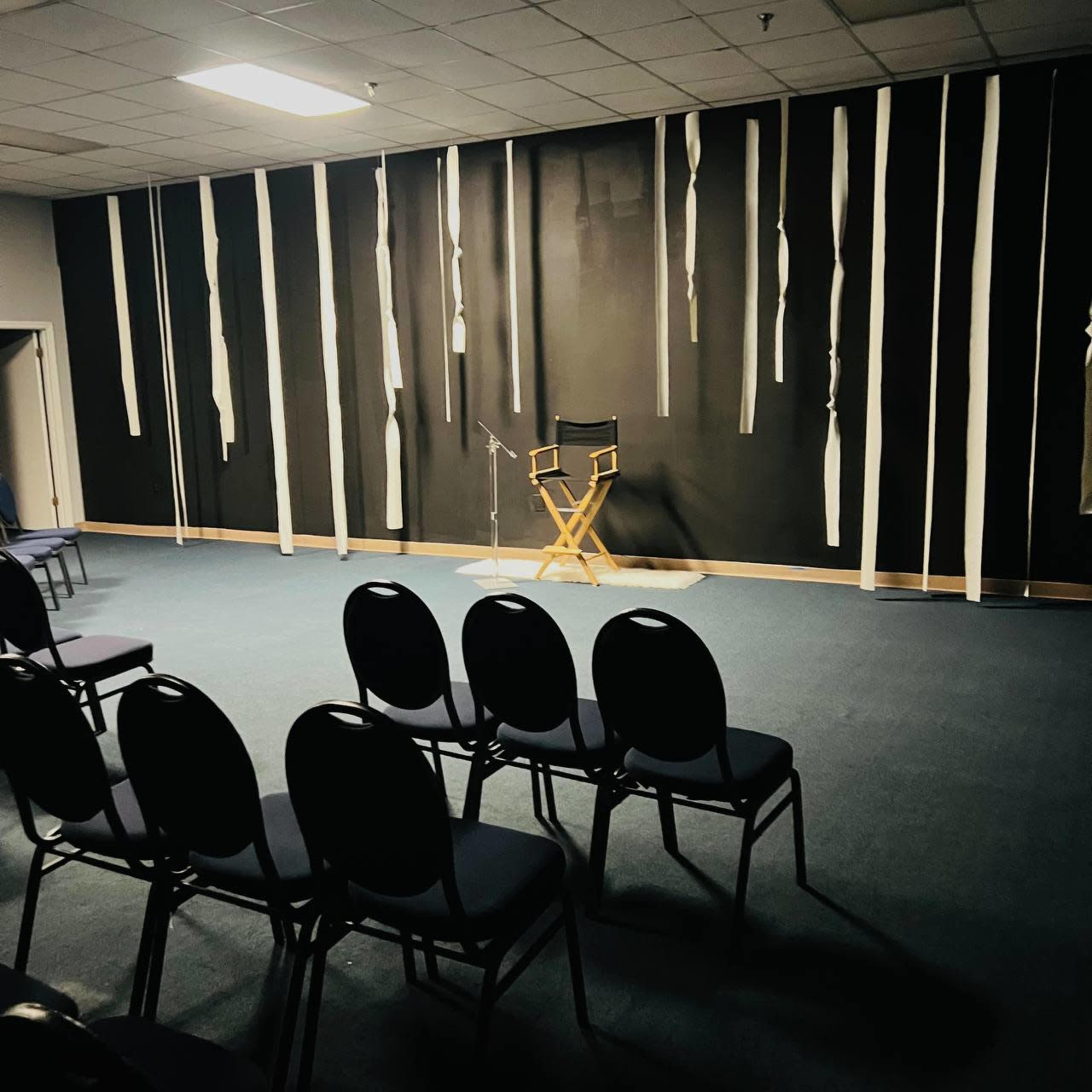 The image shows a sparse room with several rows of empty chairs facing a stage featuring a tall director's chair and a microphone against a dark wall with hanging strips of material.