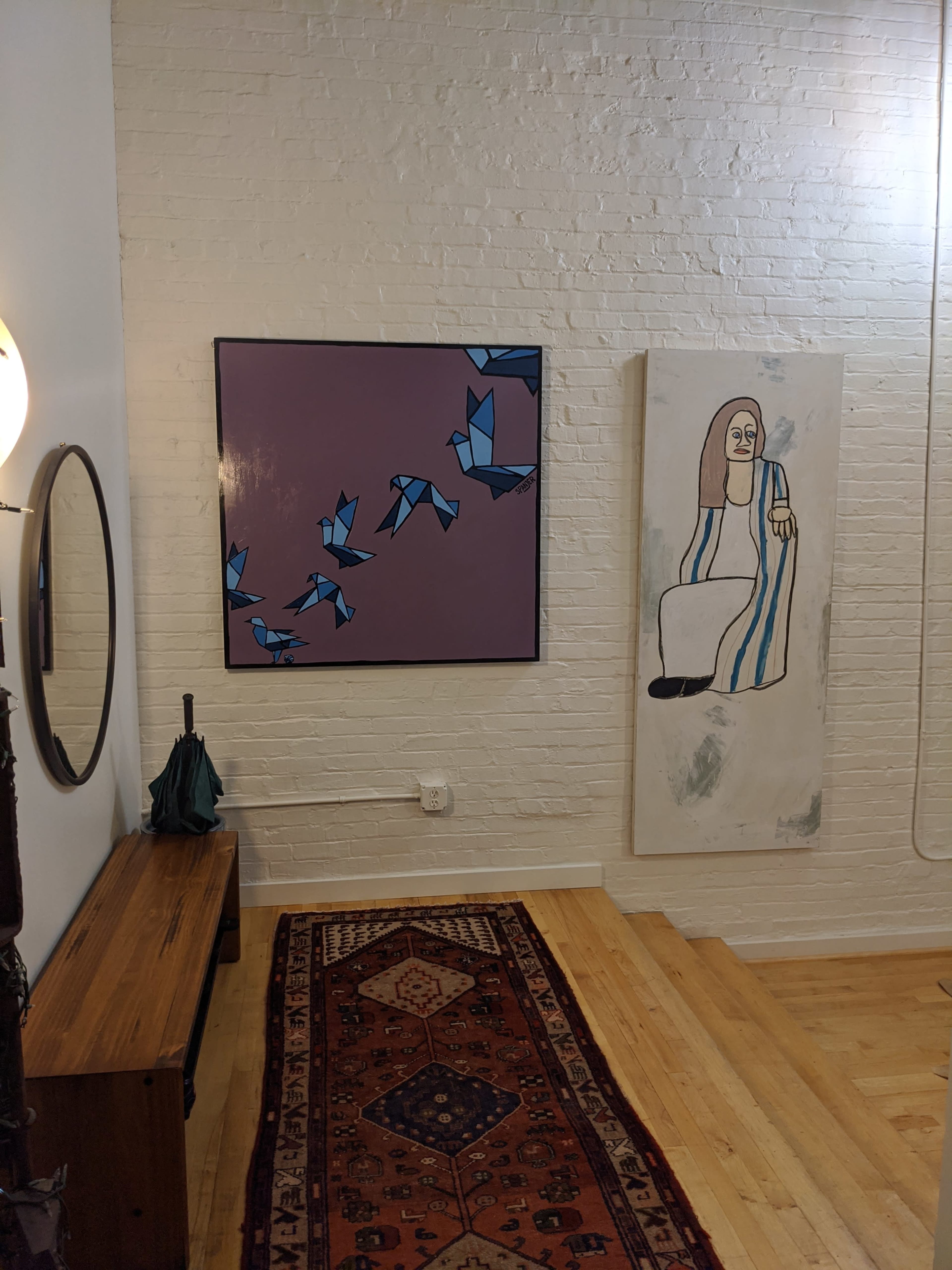 The image shows a corner of a room featuring two abstract paintings on the white brick wall, a round mirror, a wooden bench, an umbrella, and a patterned area rug.