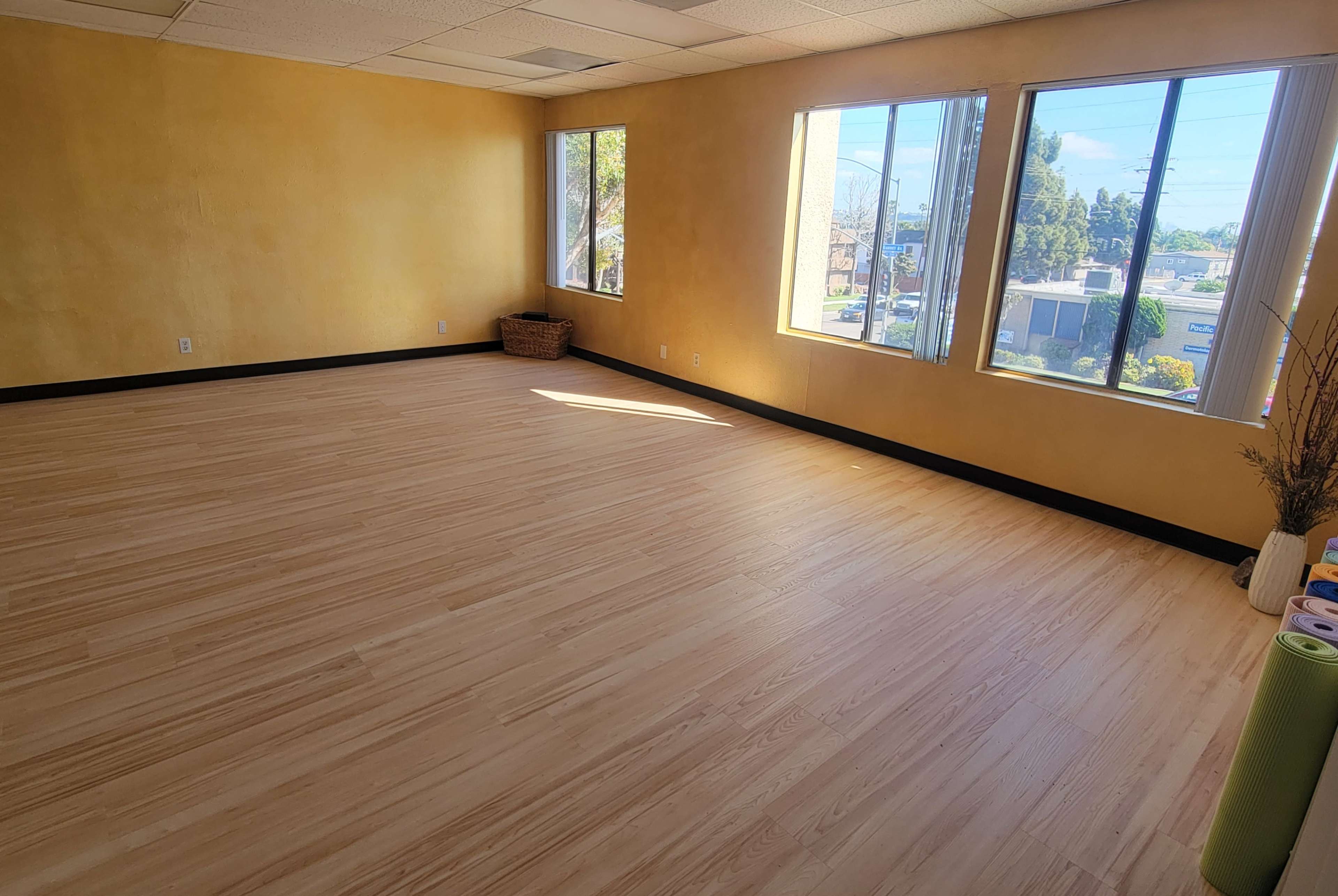 The image shows a vacant room with wooden flooring and large windows, illuminated by natural light, featuring yellow-painted walls.