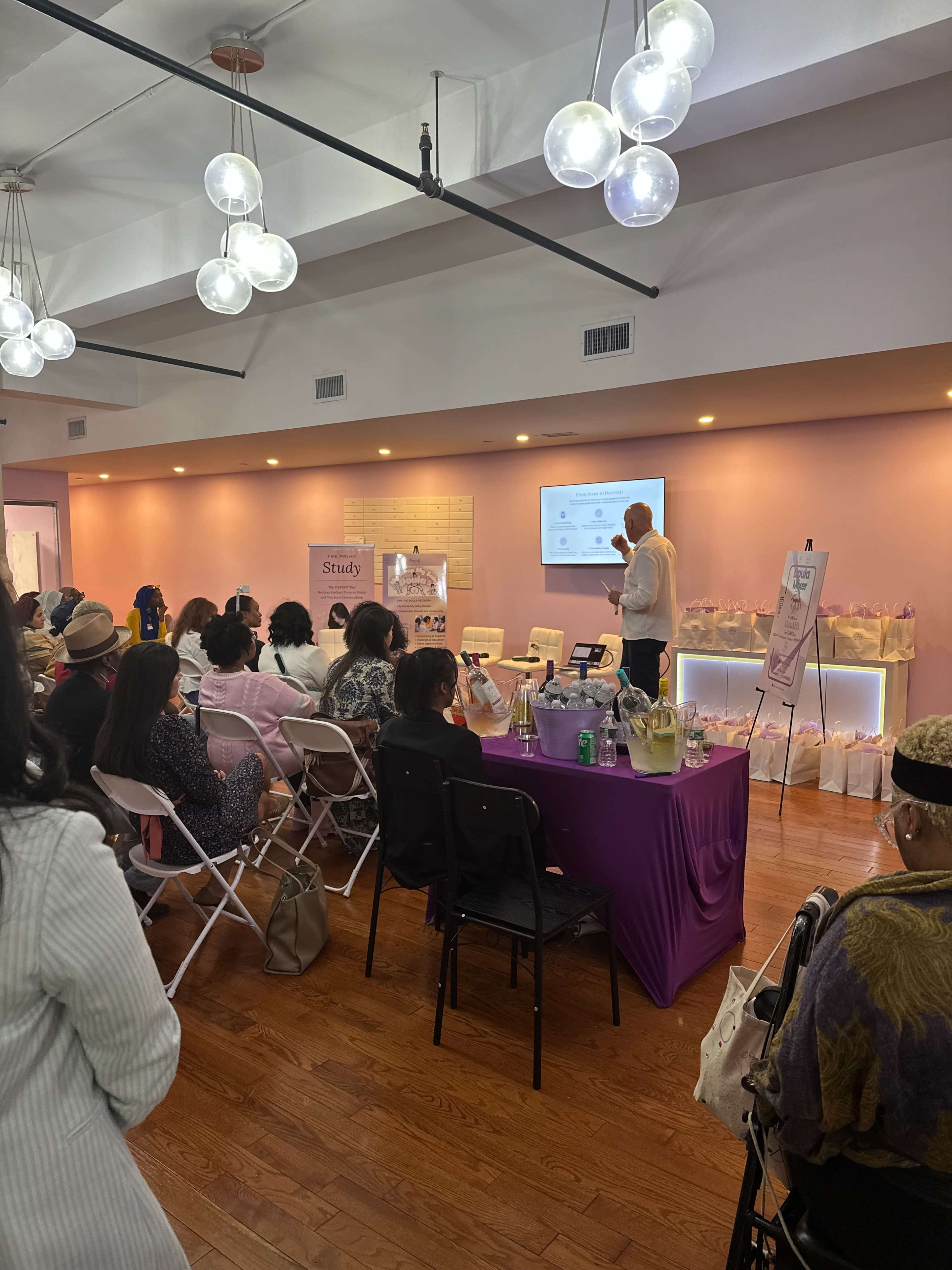 A speaker presents to an audience seated in a brightly lit room decorated with pastel colors and a display of products on tables.